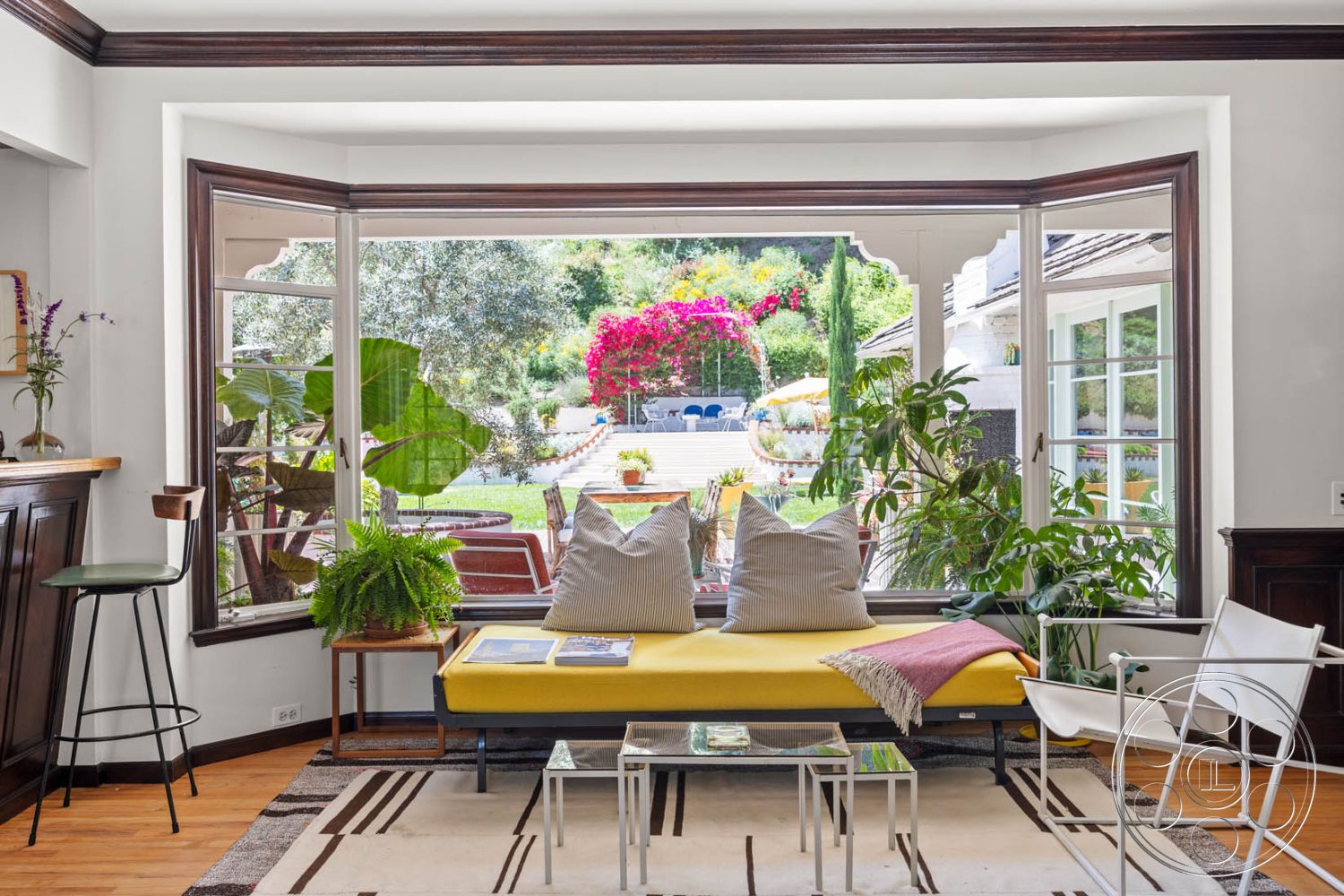 English 60 - interior white painted walls, exterior landscaped garden view, interior glass coffee table, interior hardwood floor living room, architectural style Mid-Century Modern, interior potted plants for decor, interior bay windows with wood trim, interior yellow upholstered sofa, interior striped area rug on hardwood floor, interior modern accent chair, exterior patio area with seating, exterior vibrant flower beds, architectural detail wood bar with cabinetry, structural element exposed wood ceiling beams, room function relaxing living space