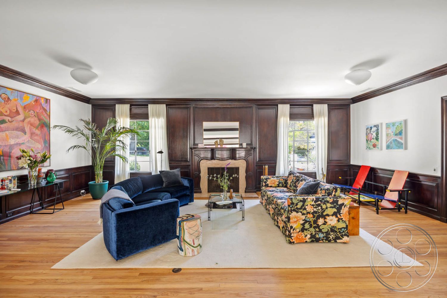 English 60 - hardwood flooring, artistic wall decor, modern light fixtures, neutral area rug, floral patterned sofa, decorative coffee table, dark wood paneling walls, interior living room, large windows with sheer curtains, potted indoor plant, upholstered navy blue couch, exposed wood mantlepiece, colorful accent chairs, elegant fireplace surround