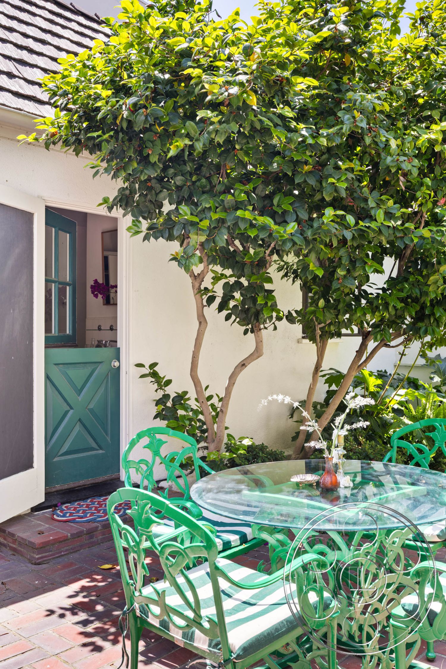 English 60 - exterior, white stucco walls, glass dining table, metal garden furniture, orange tree, potted flowers, shingle roof, lush greenery setting, landscaped garden beds, brick patio flooring, green painted wrought iron chairs, green door with diagonal paneling, colorful patterned doormat