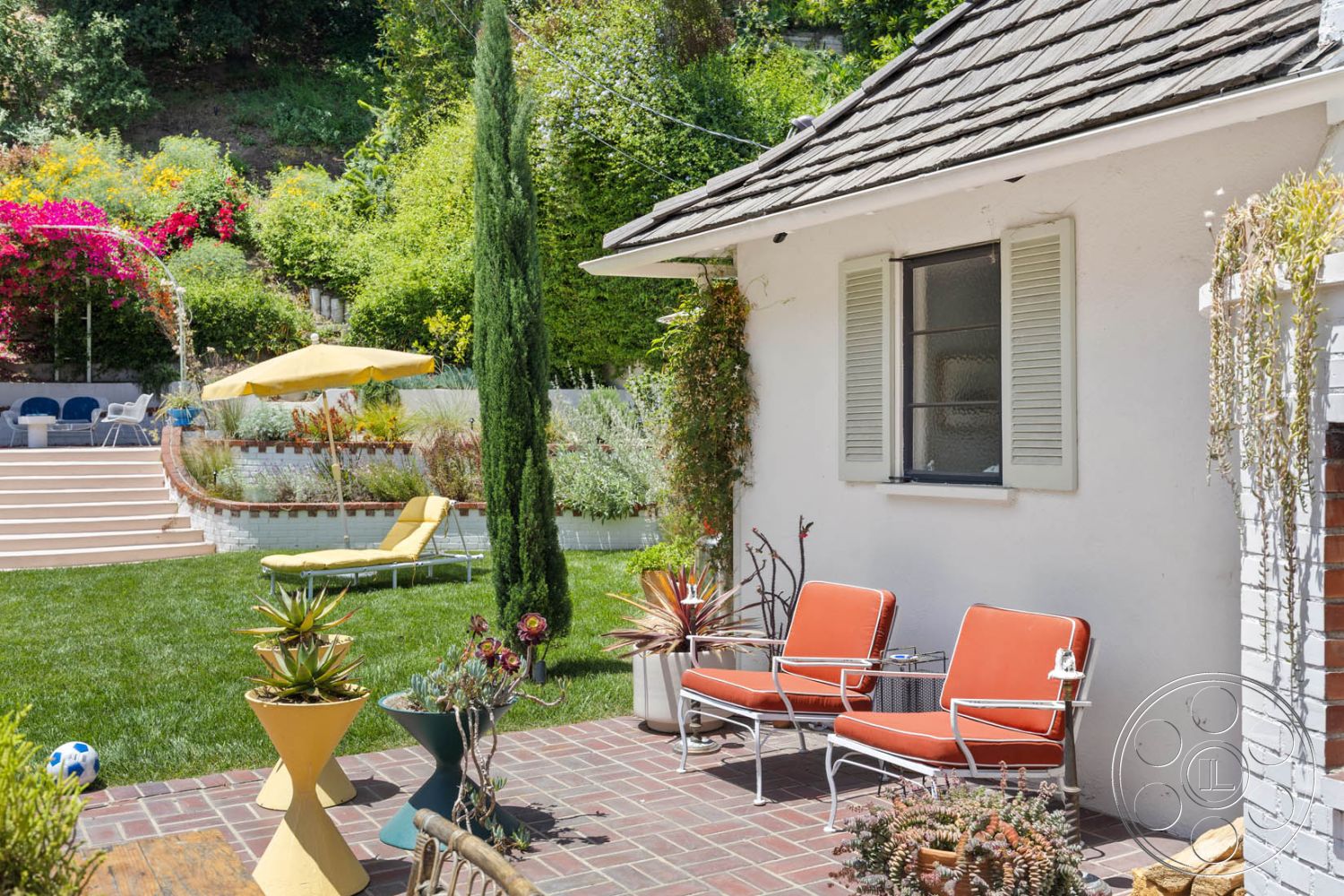 English 60 - landscaped garden, modern outdoor decor, green grass lawn, lush greenery backdrop, flat roof design, stucco exterior walls, casual outdoor seating area, brick patio flooring, sunny outdoor environment, residential backyard setting, wooden garden furniture, metal outdoor chair, beige lounge chair, with umbrella shade, blooming floral accents, tiered garden steps, circular potted plants, semi-circular retaining wall