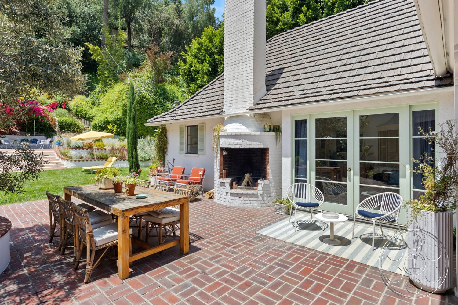 English 60 - outdoor dining area, outdoor lounge area, arched doorways, white stucco walls, wooden dining table, modern outdoor seating, lush garden setting, interior-exterior flow, shingle roof, exposed brick fireplace surround, brick patio flooring, gabled roof architecture, rattan dining chairs, lush foliage backdrop, brick wall accent, green sliding patio doors, landscape with flowering plants, hardwood garden table, textured brick chimney