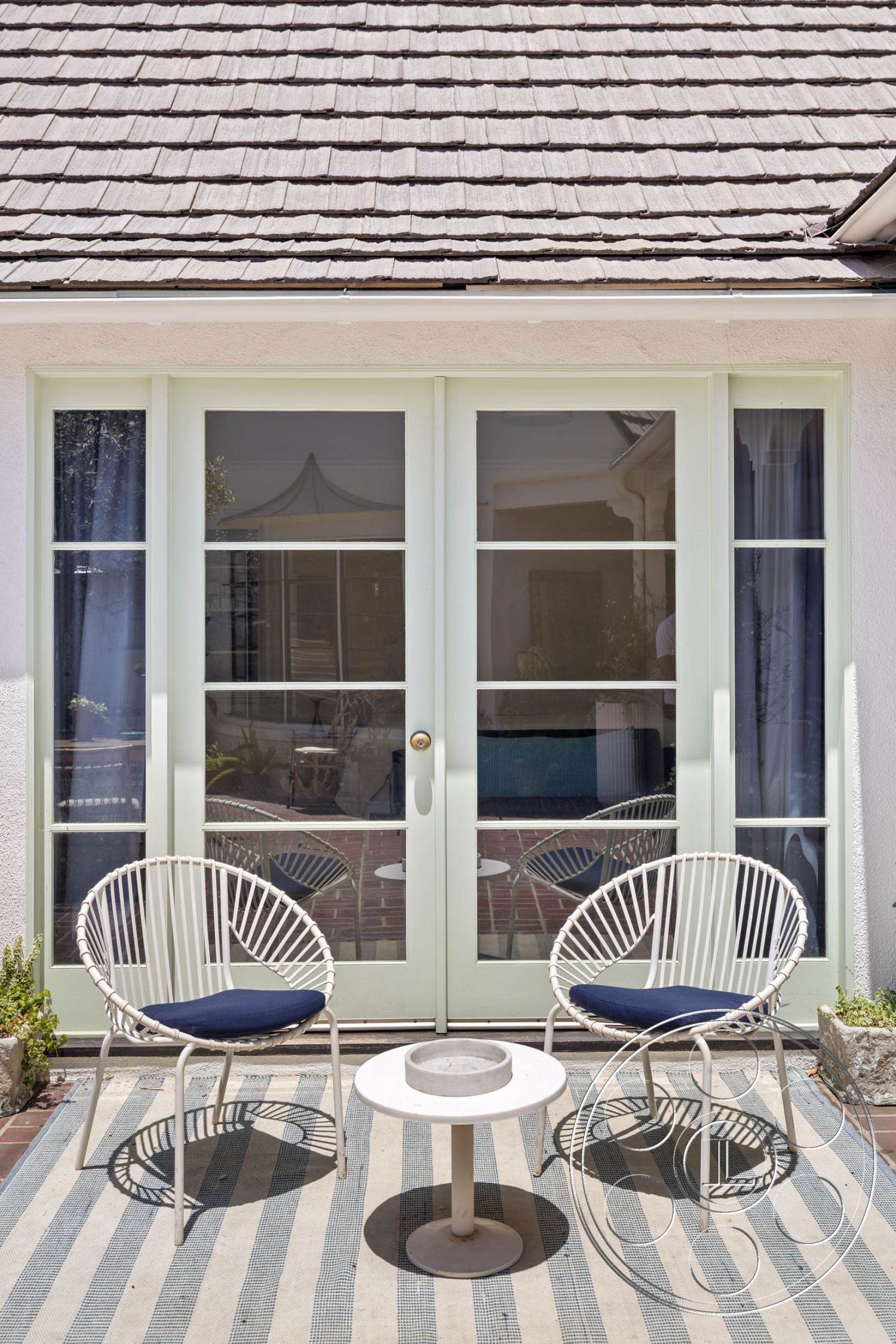 English 60 - striped outdoor rug, exterior patio area, modern patio design, brick paver flooring, casual outdoor lounge, planted greenery accents, light green glass door, white wire patio chairs, navy blue chair cushions, concrete round side table, shingle roof details, sunlit outdoor seating