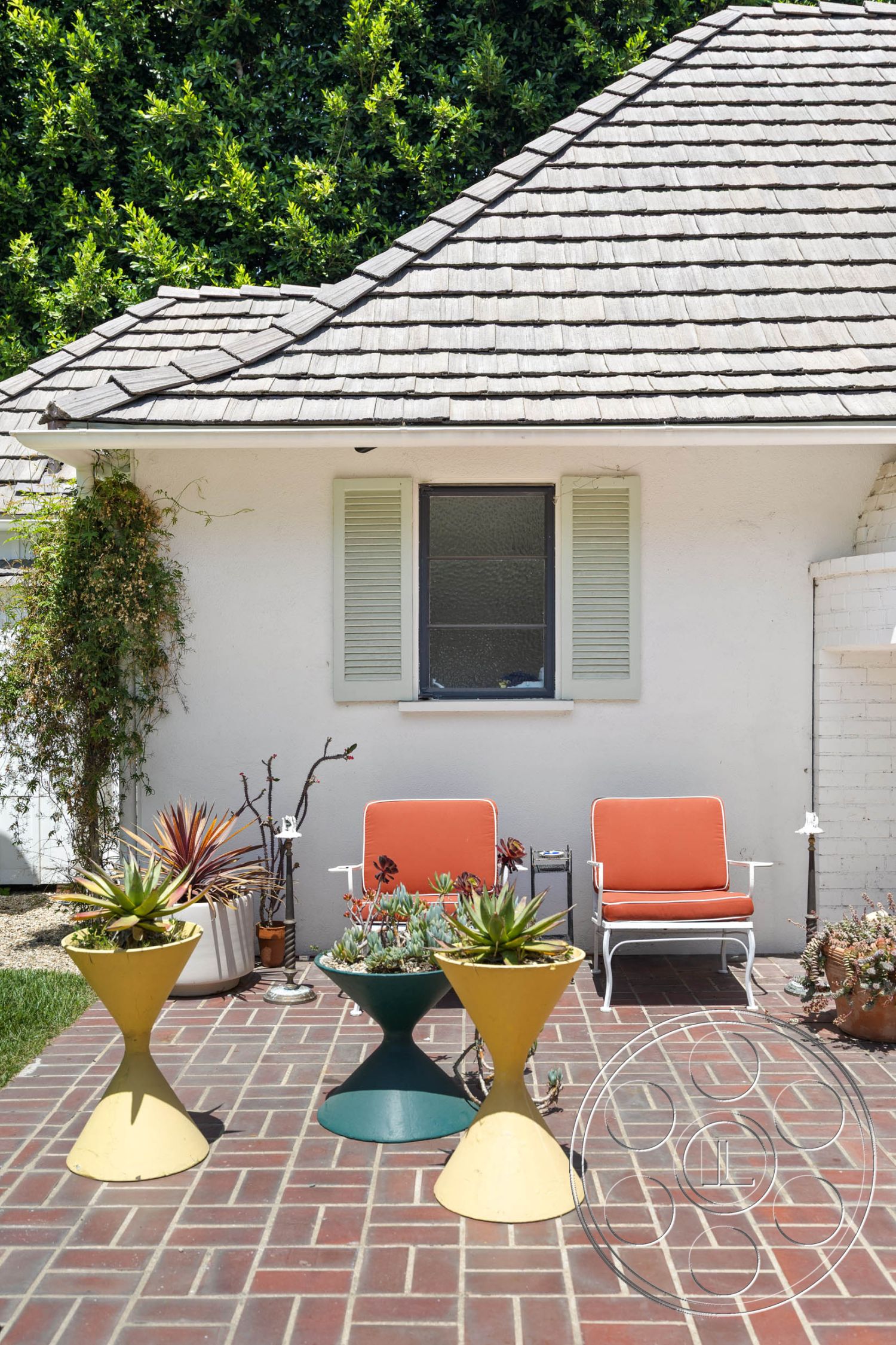 English 60 - exterior, outdoor seating area, minimalist patio design, brick patio flooring, landscaped greenery backdrop, painted stucco walls, flat roof with shingles, green window shutters, succulent plant pots, mid-century modern planters, orange outdoor chairs