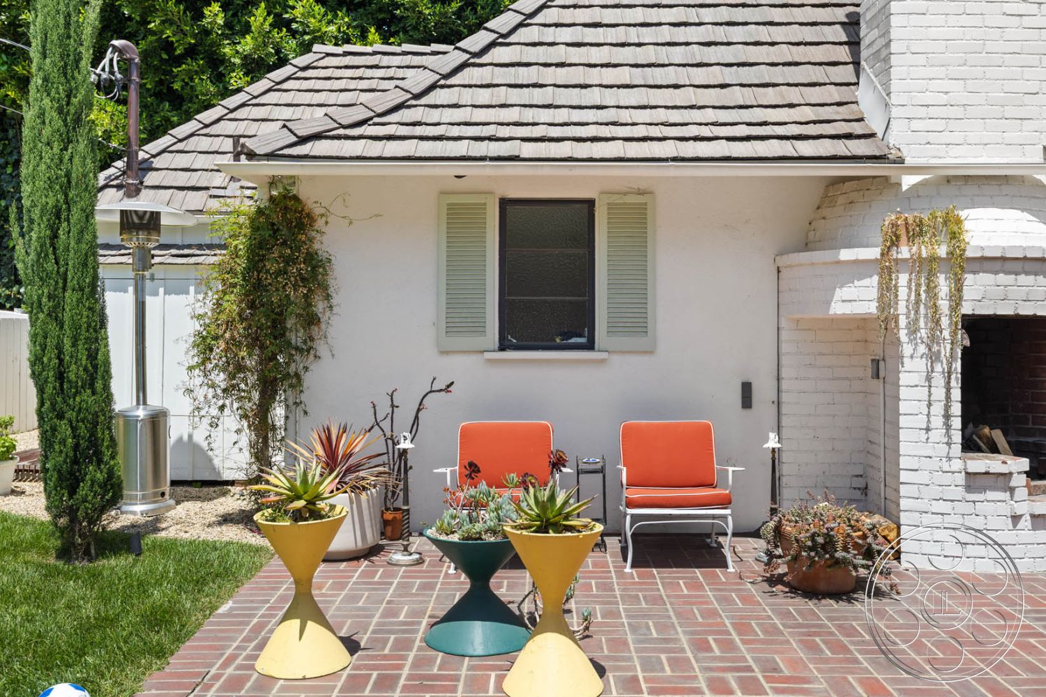 English 60 - outdoor seating area, clay tile roof, metal patio heater, mid-century modern architectural style, urban backyard setting, grassy lawn area, red brick patio flooring, modern outdoor lounge chairs, white brick exterior wall, yellow and turquoise modern planters, green succulents and potted plants, double-hung window with green shutters, exterior fireplace with brick surround, landscaped garden elements