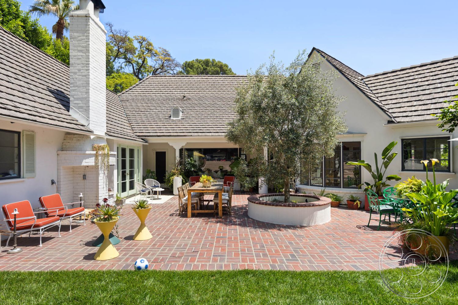 English 60 - exterior landscaped garden, exterior glass sliding doors, exterior wooden dining table, exterior brick patio flooring, exterior manicured lawn, exterior outdoor seating area, exterior potted plants, exterior residential courtyard, exterior raised circular garden bed, exterior white painted chimney, exterior sloped roof with asphalt shingles, exterior metal outdoor chairs, exterior olive tree, exterior green accent furniture, exterior suburban home setting