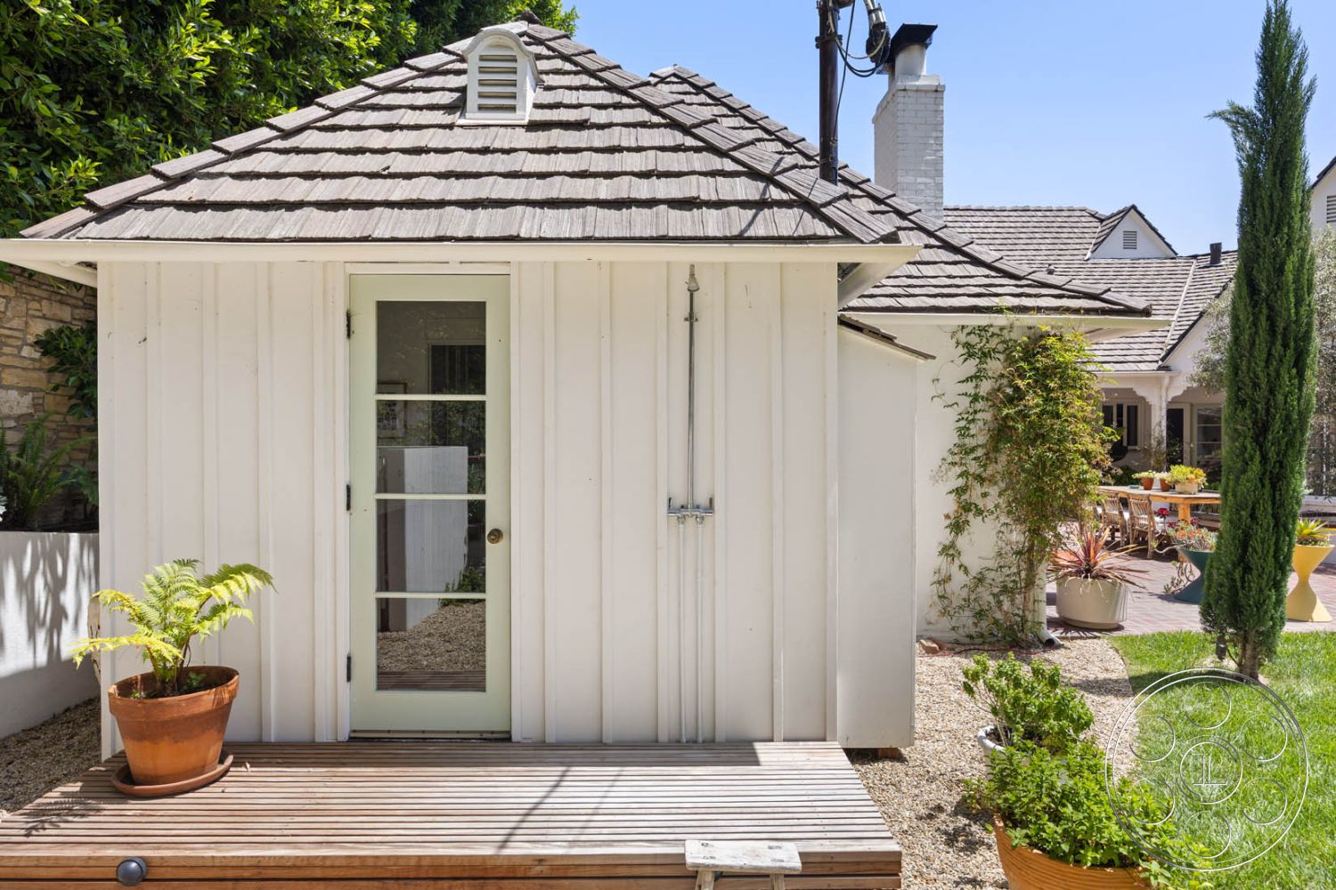 English 60 - exterior, outdoor seating area, modern outdoor furniture, gravel landscaping, urban garden setting, natural sunlight exposure, lush greenery surrounds, wood shake roof, white vertical siding, glass-paneled door, wooden deck entryway, potted fern on deck, stone wall accent, brick patio depth, cypress tree accents, gated backyard access