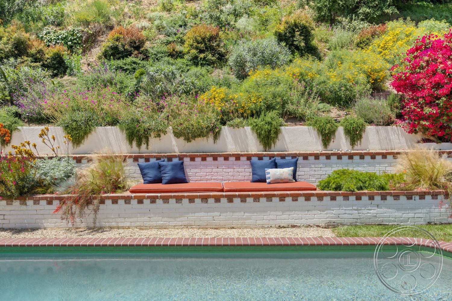 English 60 - exterior, flagstone walkway, concrete patio border, brick pool border, blue outdoor cushions, white painted brick walls, brick seating wall, landscaped garden with wildflowers, outdoor bench with cushions, textured concrete retaining wall, greenery backdrop with blooming flowers, sunny backyard with pool area, surrounding hillside landscaping
