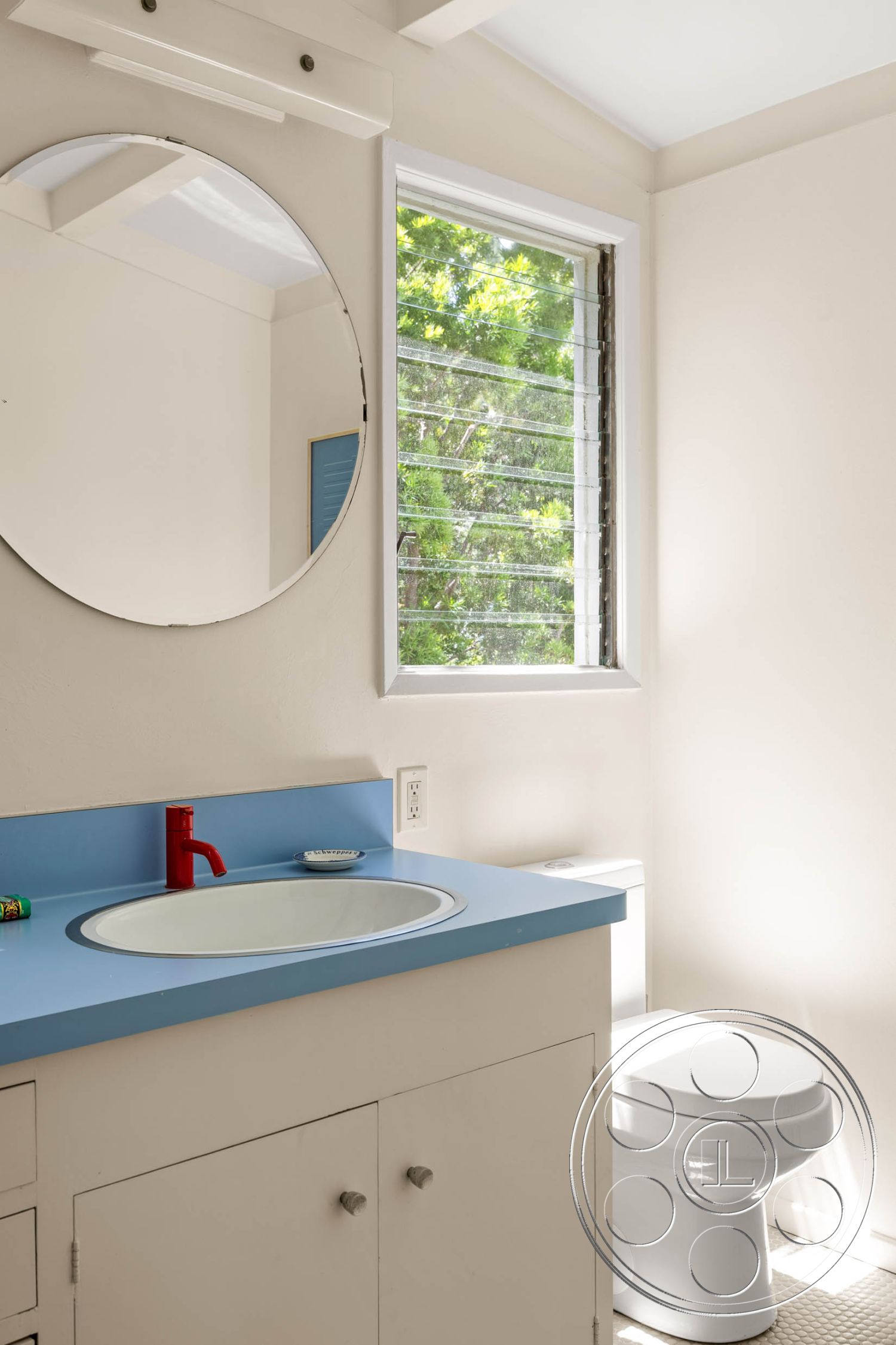 English 60 - round mirror, natural light from window, simple geometric shapes, light-colored cabinetry, modern faucet design, minimalist bathroom design, interior bathroom, white painted walls, functional bathroom layout, blue laminate countertop, circular sink, red faucet fixtures, closed toilet bowl, window with louvered glass panes, soft flooring