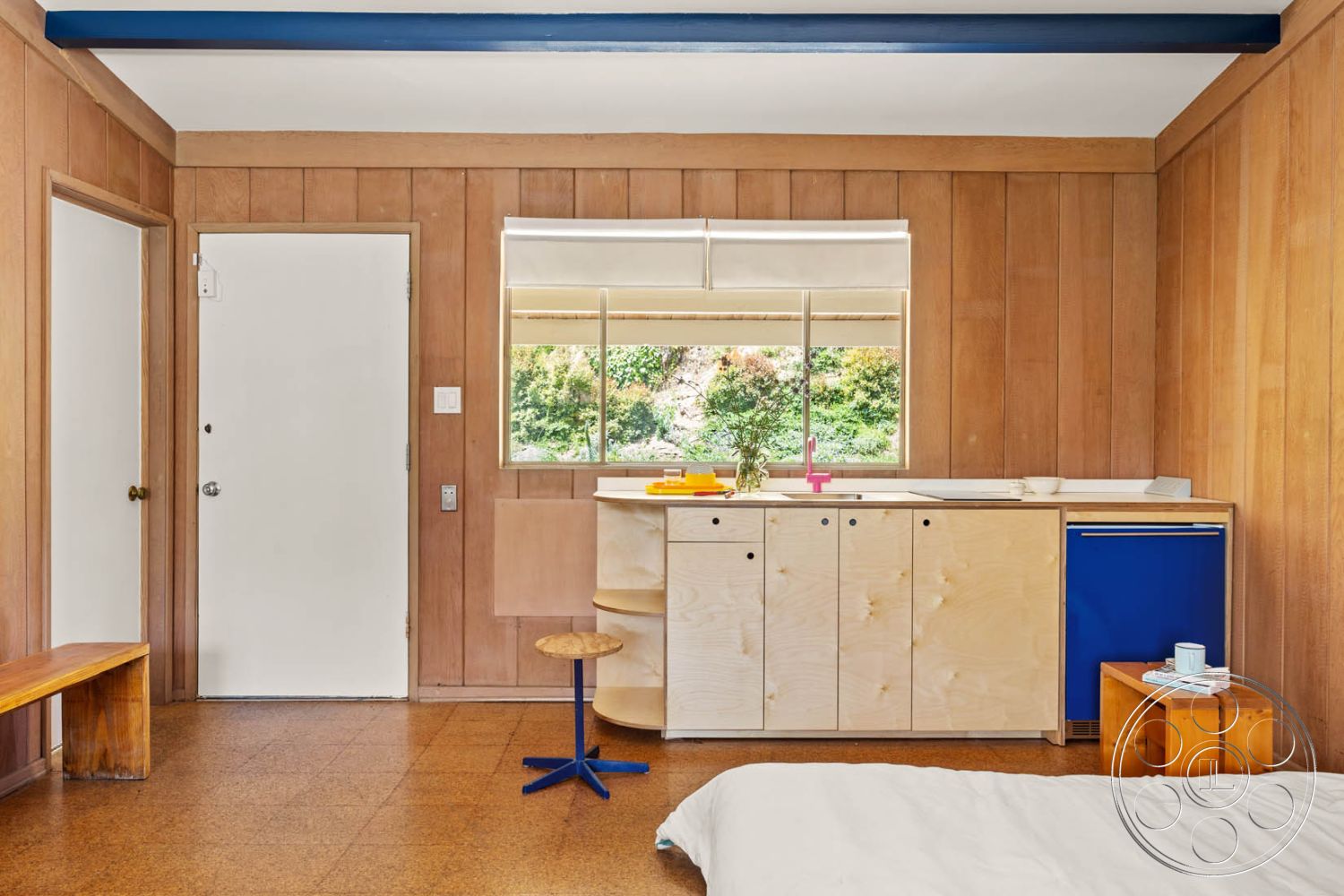 English 60 - neutral color palette, natural light from large window, modern minimalist aesthetic, open floor plan layout, multi-functional living space, exposed wooden beams ceiling, interior plywood wall paneling, laminate kitchen countertop, white-painted door, window with exterior view, cork flooring in living area, minimalist plywood cabinetry, small round wooden side table, blue accent cabinetry, functional kitchenette design, small formal entryway, nature view from interior