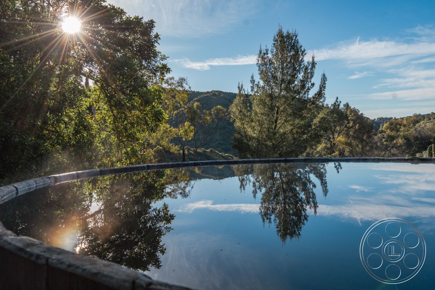Pad 90210 - sunlight filtering through trees, lush greenery backdrop, tranquil outdoor environment, exterior pool reflecting water surface, circular pool design, surrounded by natural landscaping, mountainous forest setting, wooden pool border, serene reflection in water