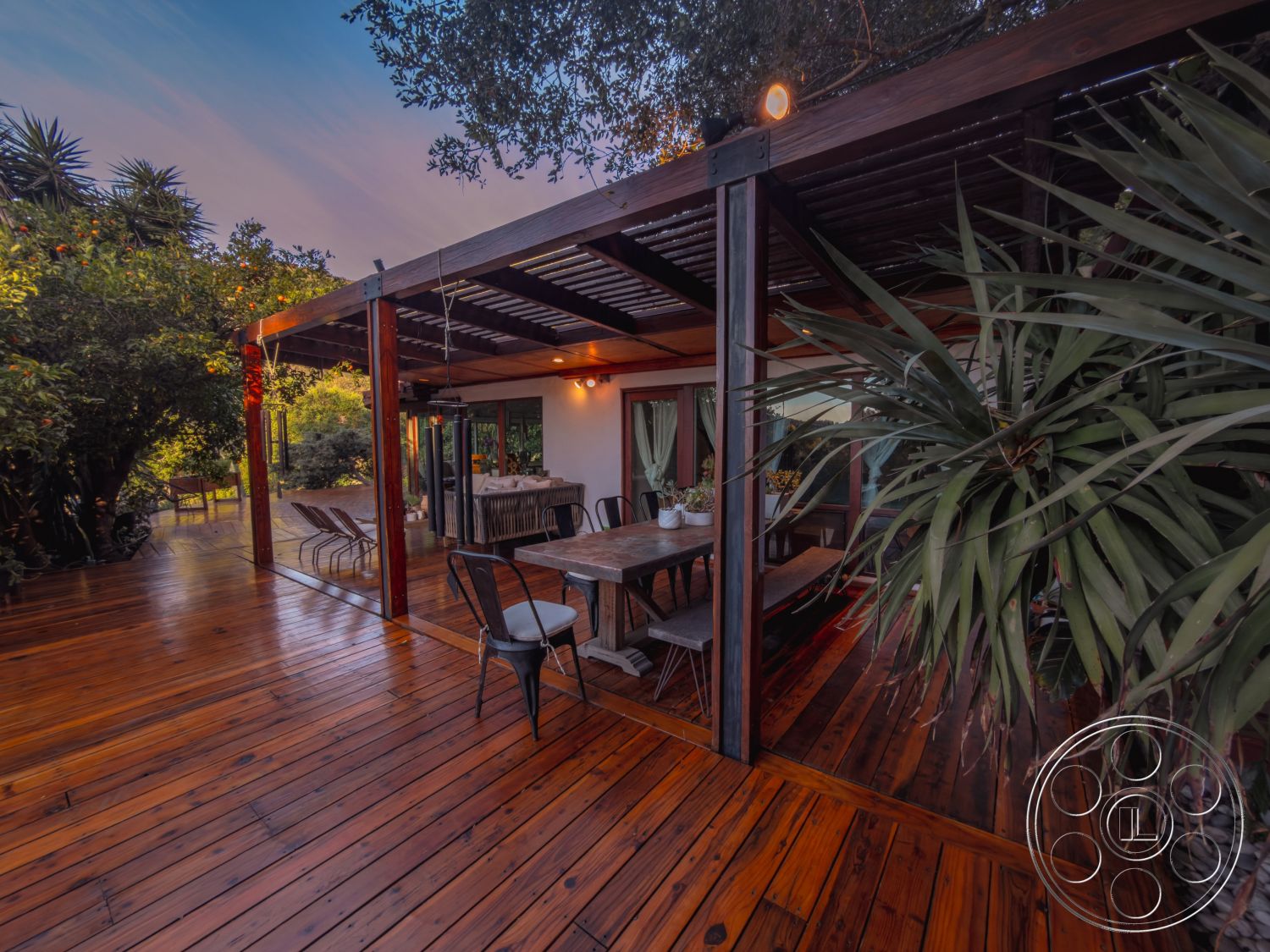 Pad 90210 - decorative potted plants, natural light through large windows, exterior wooden deck flooring, open floor plan living space, rustic outdoor furniture, surrounding tropical vegetation, outdoor dining area with wooden table, exposed wooden beam pergola, panelled glass door entry, modern metal chair seating, dim ambient lighting fixtures, wooden deck with polished finish, green leafy landscaping, sunset lighting through foliage