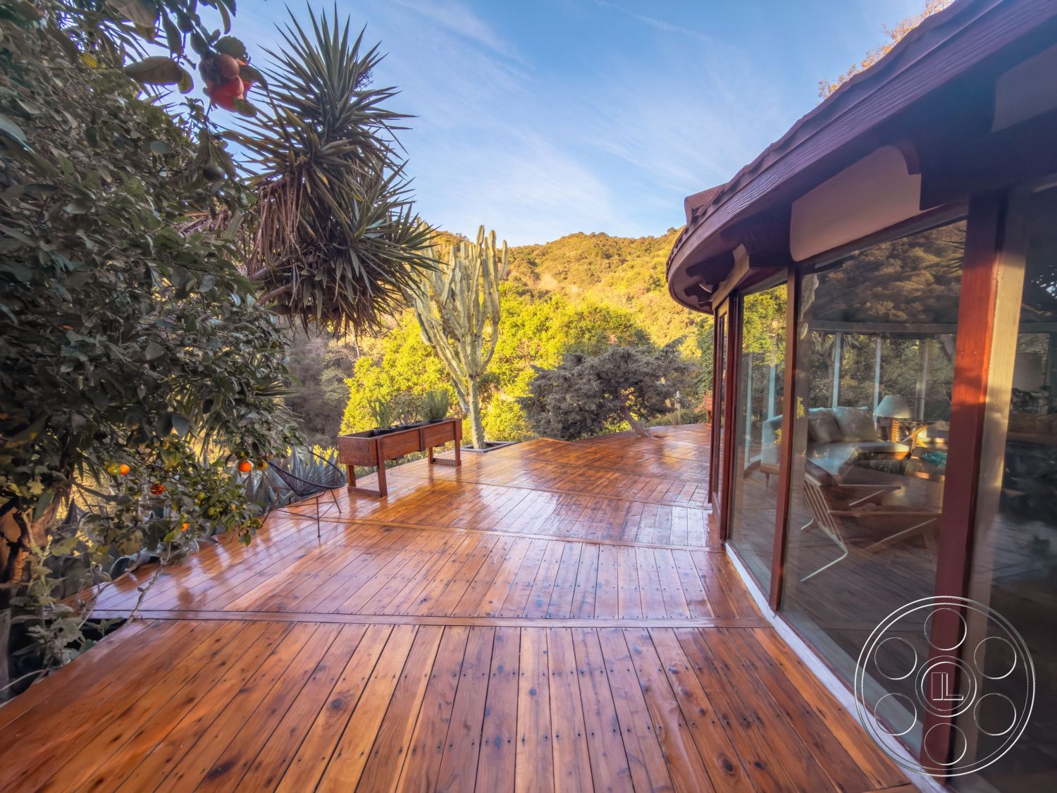 Pad 90210 - wooden bench seating, open-air living space, contemporary architectural style, natural light-filled space, exterior wooden deck, floor-to-ceiling glass windows, rustic outdoor setting, surrounding lush foliage, private garden area, panoramic mountain view, sustainable outdoor materials, cedar wood decking, curved deck design, integrated nature landscaping, desert plant accents, hillside location deck