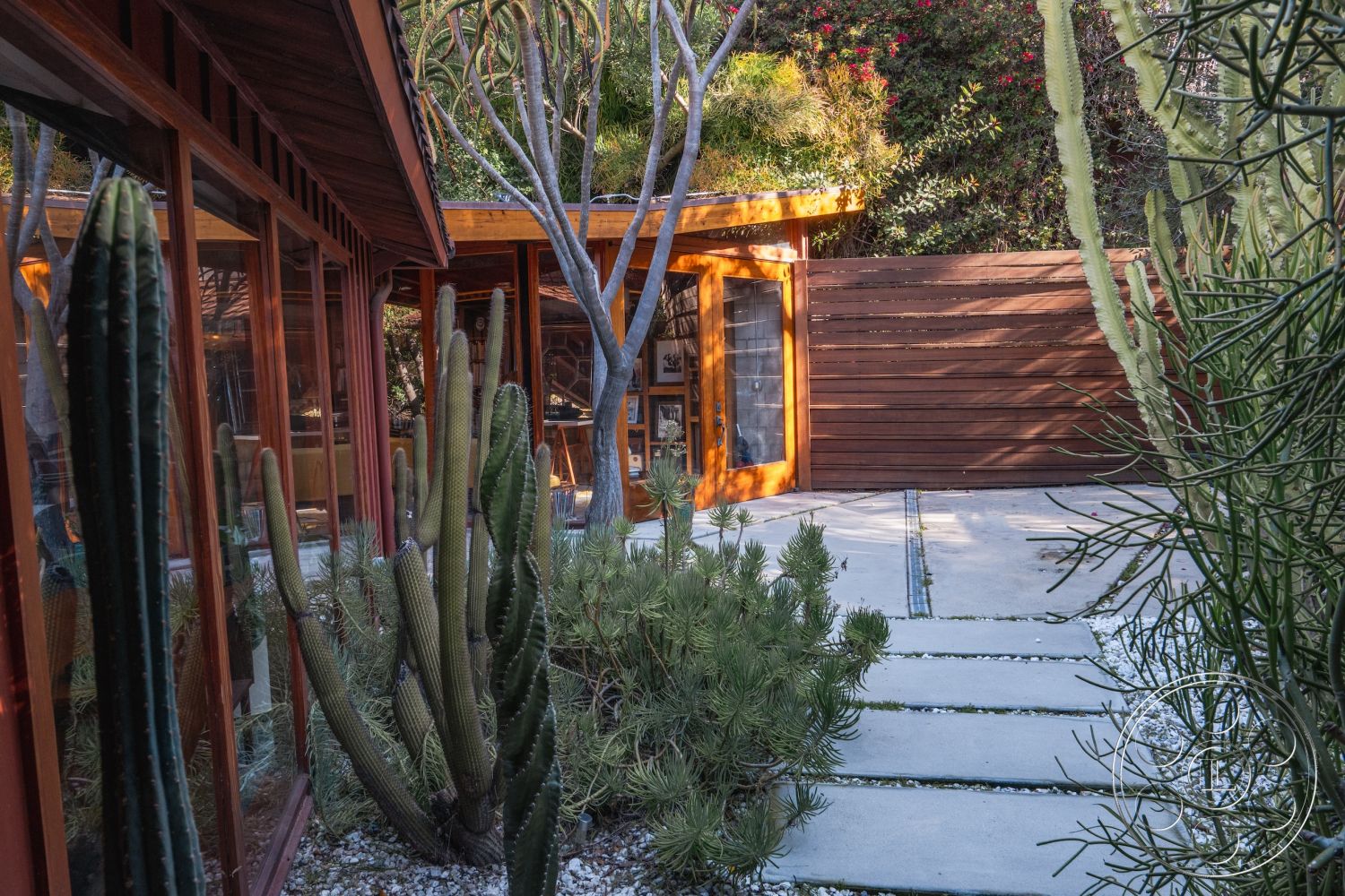 Pad 90210 - natural stone accents, exposed wooden beams, open-air courtyard, sustainable building materials, minimalist outdoor space, modern architectural design, rectangular concrete stepping stones, exterior wooden wall cladding, landscaped desert garden, contemporary outdoor living area, eco-friendly landscape design, exterior glass panel walls, concrete walkway pavers, textured gravel ground cover, native desert plants, vibrant flora accenting architecture, sunlight streaming through windows, artistic wooden entrance door, lightly stained wooden frames, harmonious indoor-outdoor flow