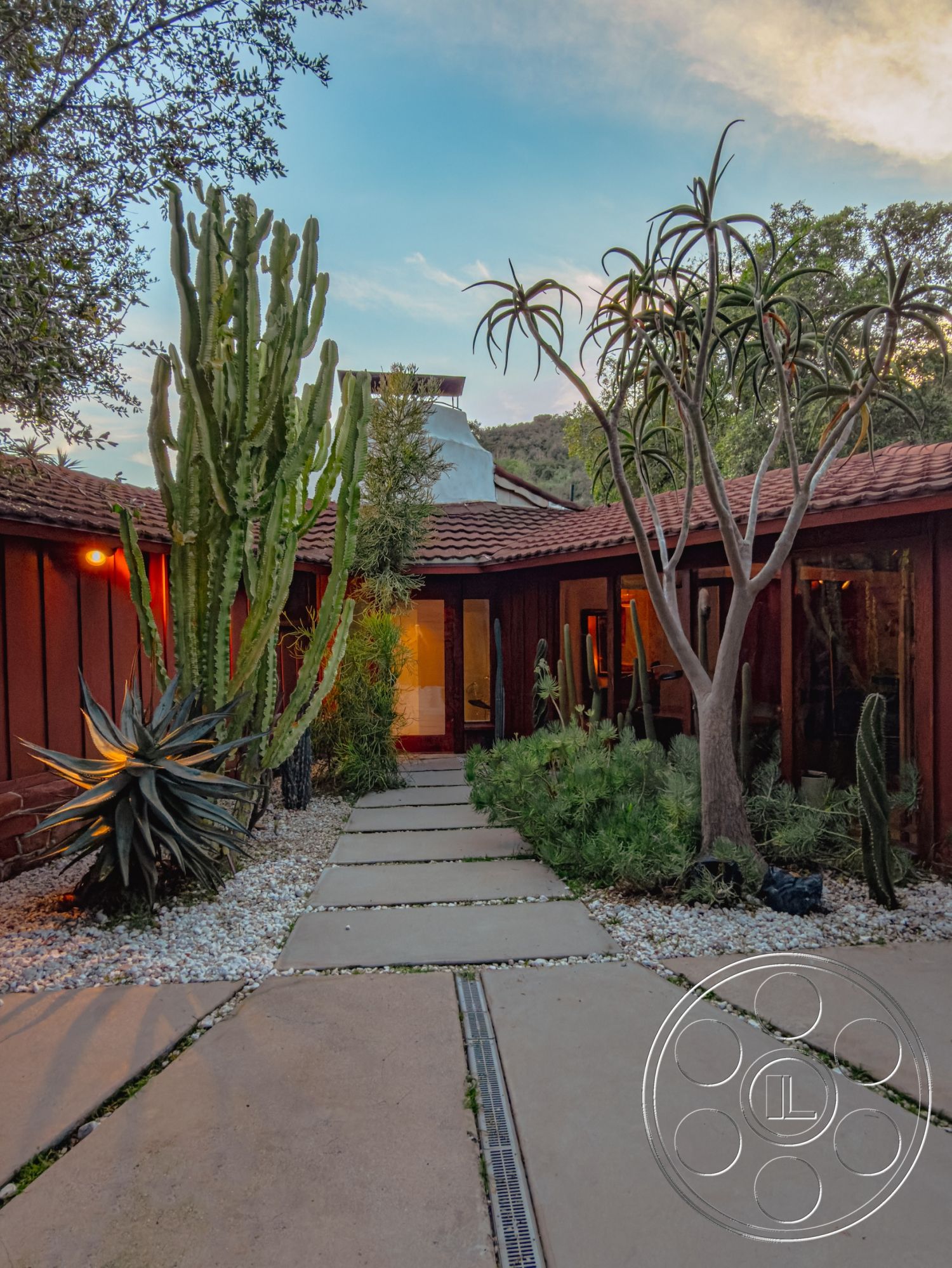 Pad 90210 - modern architectural lines, sliding glass doors, wood paneling on exterior walls, desert landscape with cacti, open courtyard layout, exterior courtyard home, concrete walkway with gravel border, red tile roofing, natural stone path pavers, greenery with succulents and shrubs, landscaped garden with gravel accents, sunset lighting ambiance, metal drainage channel, surrounding mountainous backdrop