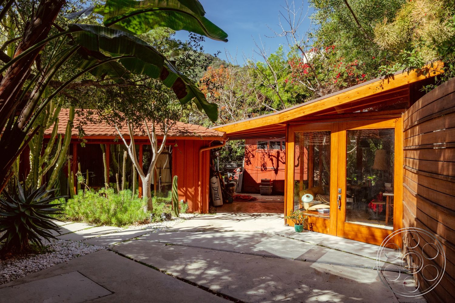 Pad 90210 - indoor-outdoor flow, exposed wood beams, inviting entryway, open-air courtyard, lush tropical landscaping, terracotta tile roof, flagstone path, concrete walkways, wooden privacy fence, concrete patio flooring, mid-century modern architectural style, urban garden setting, modern outdoor seating area, sunlit outdoor space, sloped roof architecture, natural stone garden beds, landscaped garden areas, exterior wooden door with large glass panels, red stucco exterior walls, potted succulents and cacti