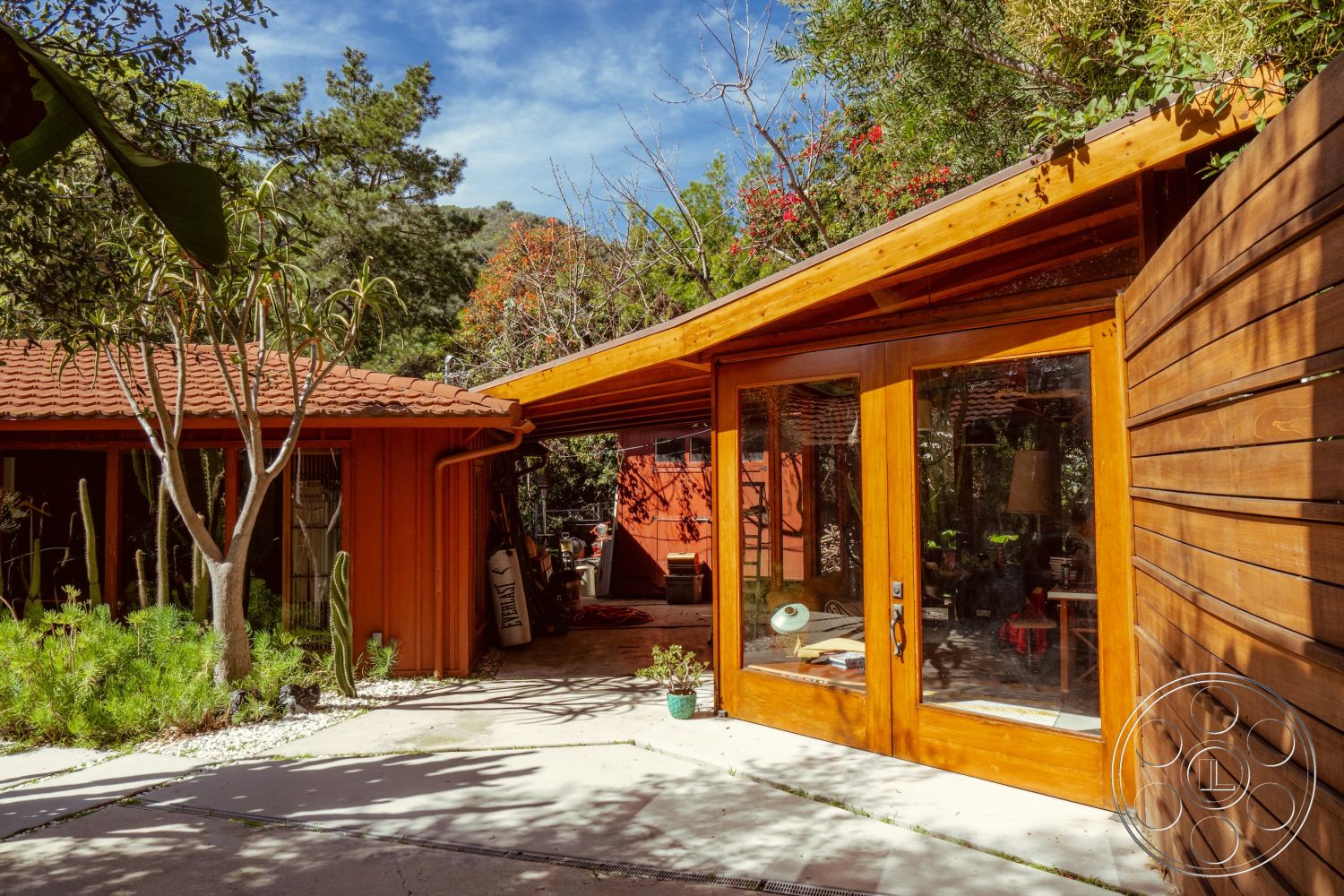 Pad 90210 - rustic charm, private outdoor space, mountain backdrop, red tile roof, sunny exterior, concrete patio flooring, mid-century modern architectural style, outdoor garden area, wood beam roof overhang, floor-to-ceiling glass windows, interior greenery view, wooden exterior doors, orange stucco walls, exposed wooden horizontal siding, natural landscaping with cacti, lush green setting, tertiary structural elements, sloped rooflines, aromatic garden plants