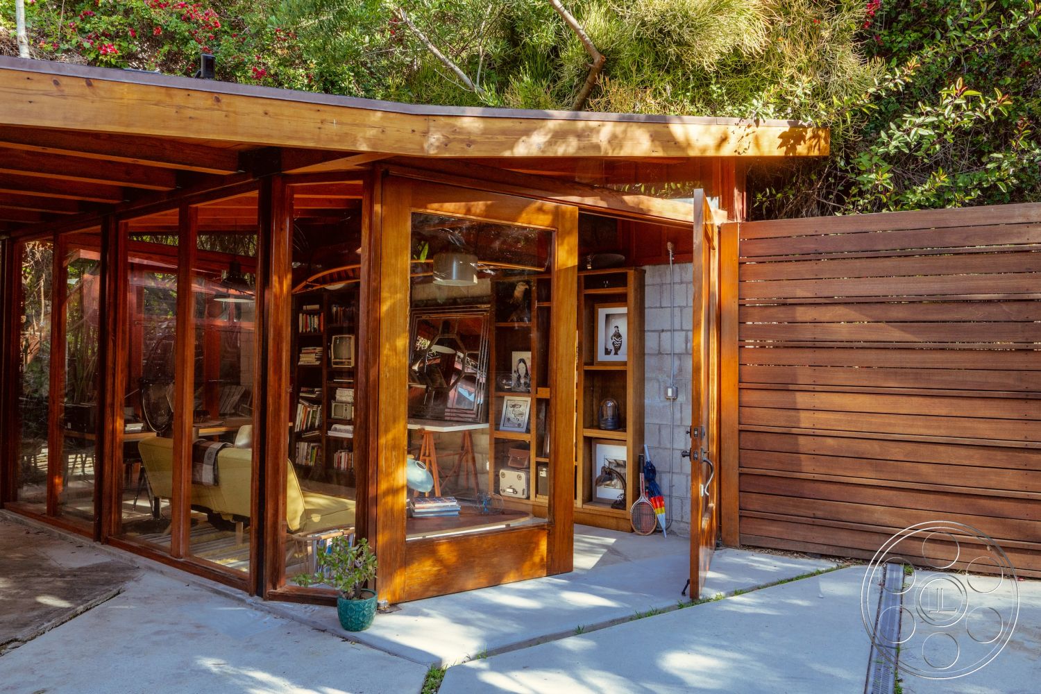 Pad 90210 - contemporary outdoor space, curved roof design, concrete patio flooring, exposed concrete block walls, rustic wood accents, wooden slat privacy fence, sliding glass door entrance, exterior wooden beam construction, large glass window features, open shelving interior design, landscape greenery surrounding