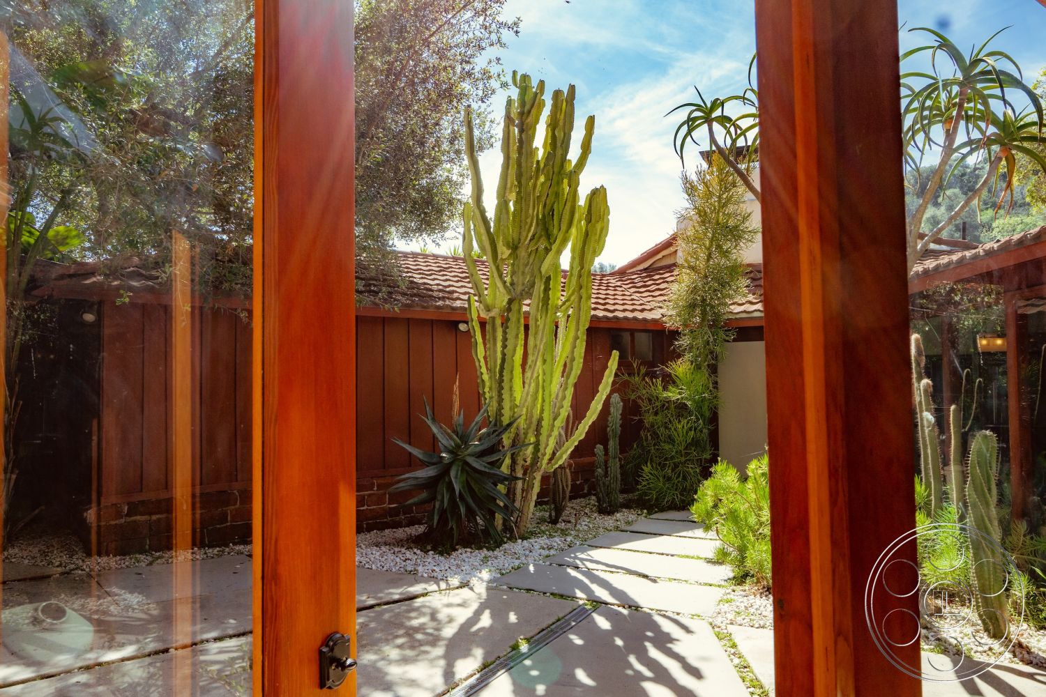 Pad 90210 - earthy color palette, red tile roof, wooden door frame, modern architectural details, glass panel doors, succulent garden plants, courtyard garden setting, natural stone gravel ground cover, desert landscaping elements, decorative stone garden path, exposed wood wall siding, outdoor paver stones, interior and exterior integration, sunlit courtyard space, vertical cactus plants, low-maintenance foliage