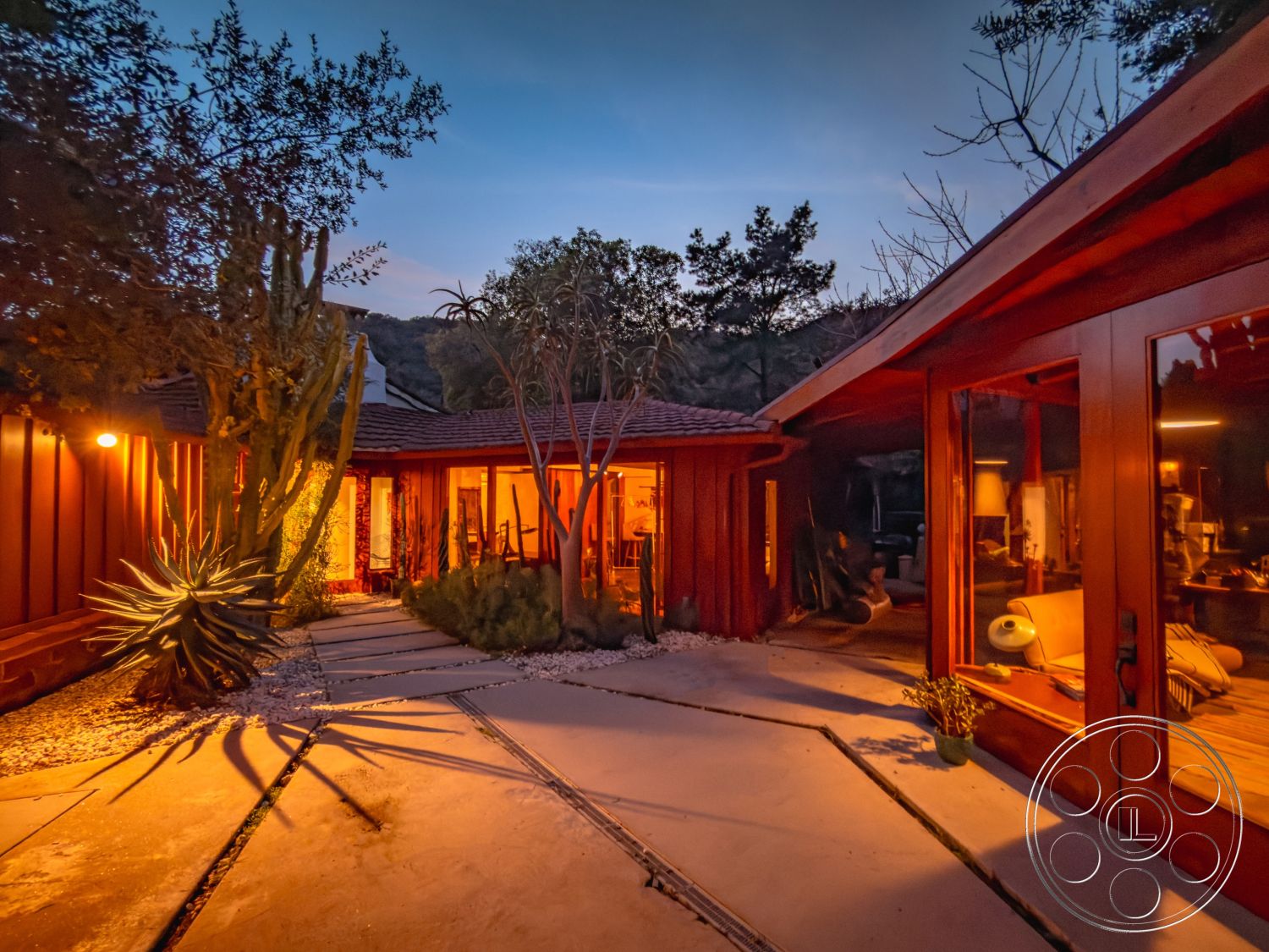 Pad 90210 - glass sliding doors, open floor plan design, modern outdoor seating area, natural stone feature wall interior, landscaped garden with native plants, soft ambient outdoor lighting, mountain landscape view, gabled roof with clay tiles, exterior patio with concrete floor pavers, exposed wood accent walls, panelled wooden siding, desert landscaping with succulents, roof overhang for shade, curved path with gravel borders, wooden deck surrounding the house, interior living room with wooden beams, wall-to-wall windows for natural light, cozy evening ambiance, outdoor integrated lighting schemes