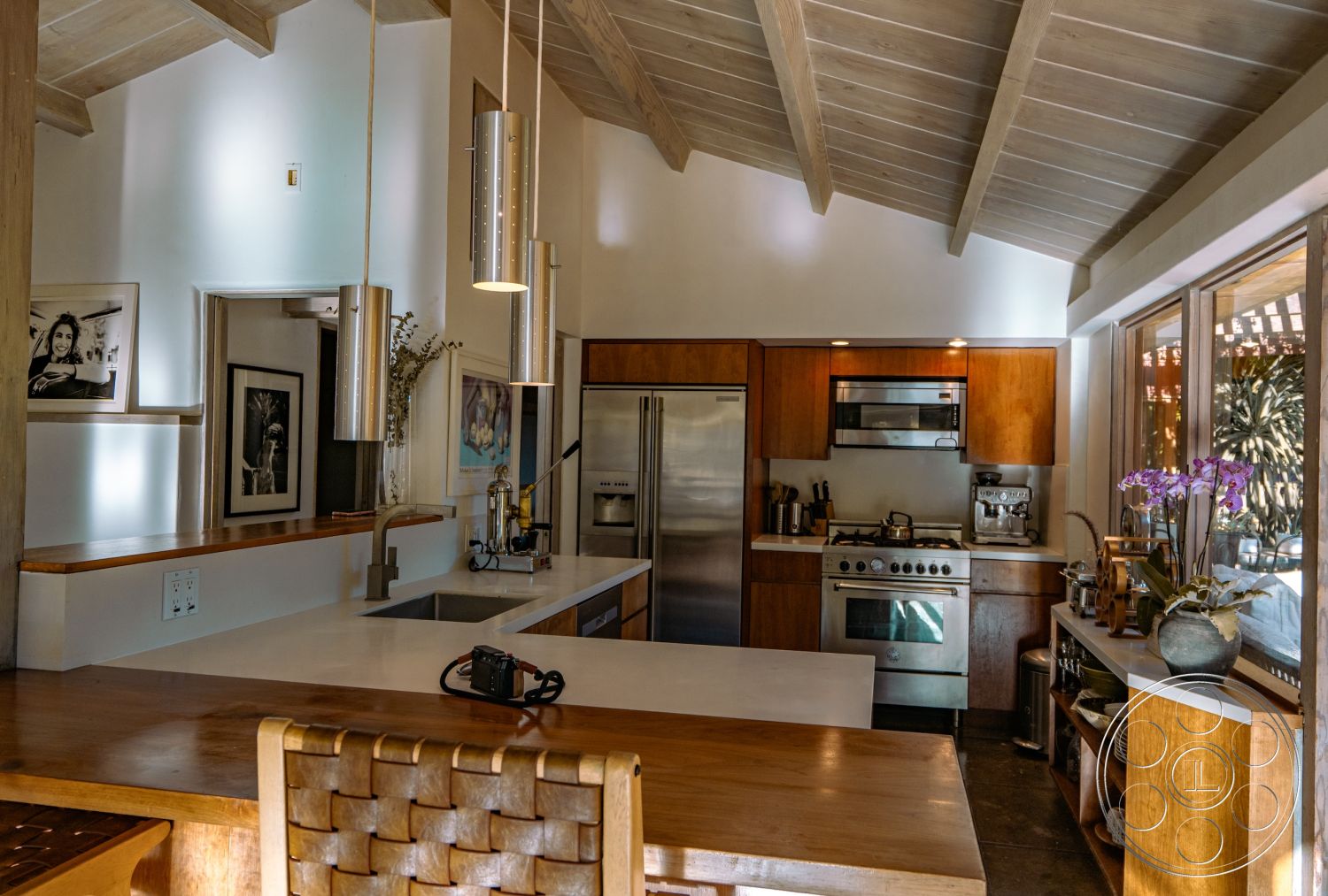 Pad 90210 - stainless steel appliances, modern art wall decor, open concept design, concrete kitchen countertop, pendant lighting fixture, natural light-filled space, minimalist kitchen design, wood cabinetry with natural finish, dark tile flooring, interior kitchen with vaulted wooden ceiling, glass window wall overlooking outdoor space, wooden bar height dining table, blooming orchids as interior decor