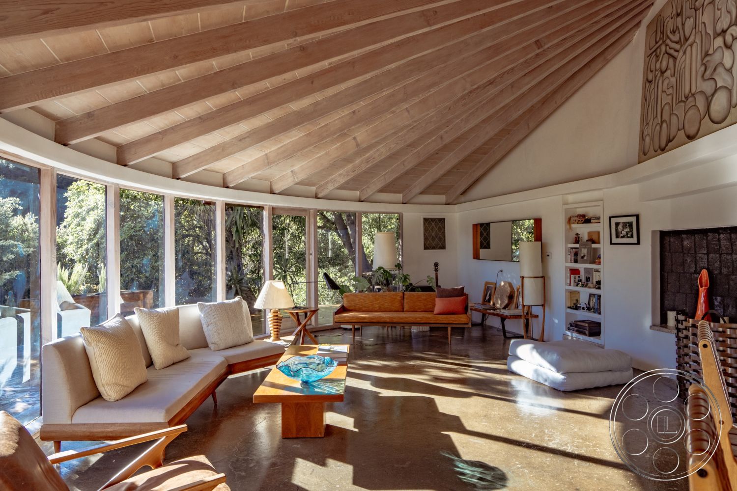 Pad 90210 - polished concrete flooring, cozy lounge area, contemporary interior design, natural wood coffee table, open-plan living space, exposed wood ceiling beams, textured plaster wall detail, floor-to-ceiling glass windows, integrated shelving units, surrounding forest view, bright interior with natural light, curved concrete interior walls, vaulted circular living room ceiling, wooden mid-century furniture, bright throw pillows, rustic stone fireplace wall