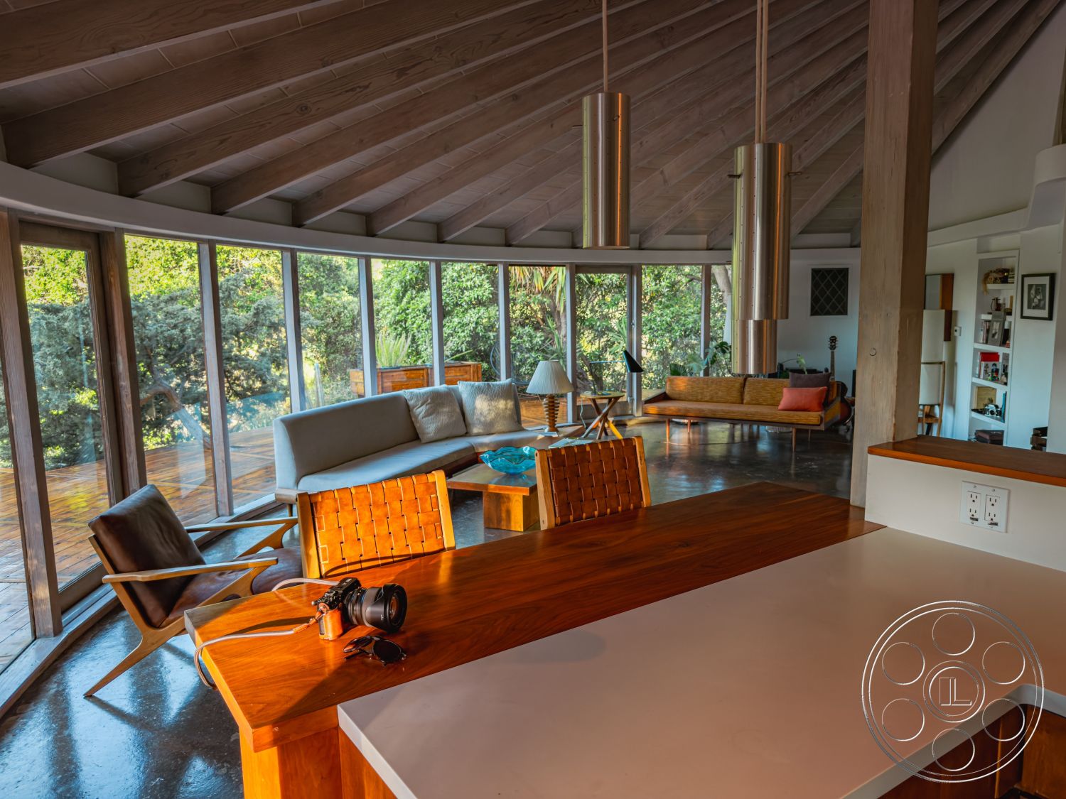 Pad 90210 - polished concrete flooring, wooden dining table, floor-to-ceiling windows, textured wall panels, cozy lounge area, urban residential setting, mid-century modern design, exposed wood ceiling beams, recessed lighting fixtures, built-in shelving units, open-plan living area, natural light-filled interior, minimalist kitchen island, curved glass wall, surrounding garden view, modern wooden chairs