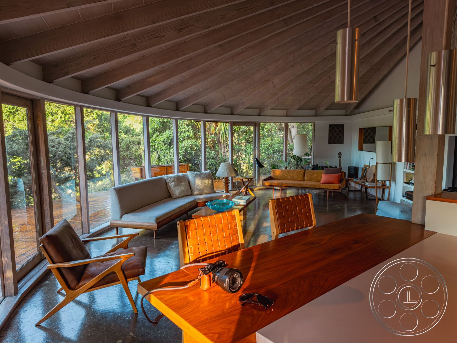 Pad 90210 - polished concrete flooring, contemporary pendant lighting, open-concept living space, textured wall paneling, seamless indoor-outdoor connection, surrounding greenery view, minimalist wooden dining table, exposed wooden support structure, large floor-to-ceiling glass windows, mid-century modern armchairs, vaulted wooden ceiling beams, modern lounge area with low-profile sofas, natural light-filled open layout, integrated modern kitchen design, wooden deck access, relaxed living room atmosphere, stylish coffee table design