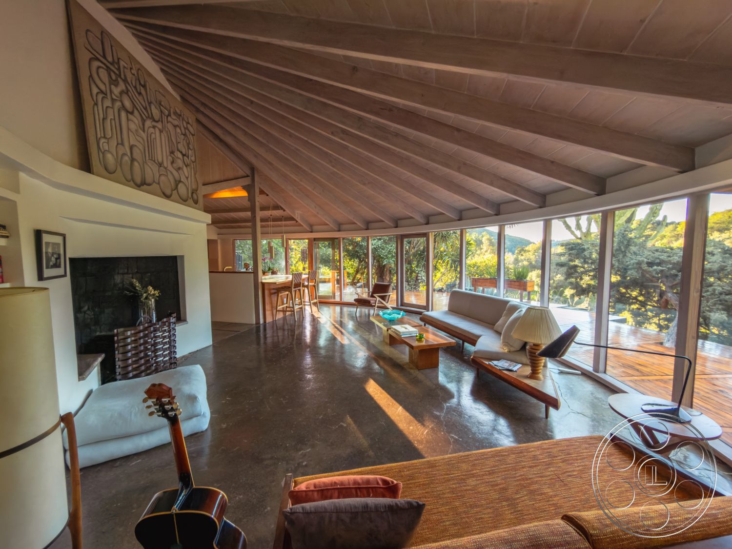 Pad 90210 - exposed wood ceiling beams, suburban backyard view, minimalist furniture design, vaulted living room ceiling, modern lounge seating area, natural light-filled interior, wood beam architectural details, open concept interior layout, large floor-to-ceiling glass windows, concrete flooring in living area, wooden dining table with bar stools, textured wall art above the fireplace, natural setting with lush greenery view, interior blending with nature, contemporary fireplace surround