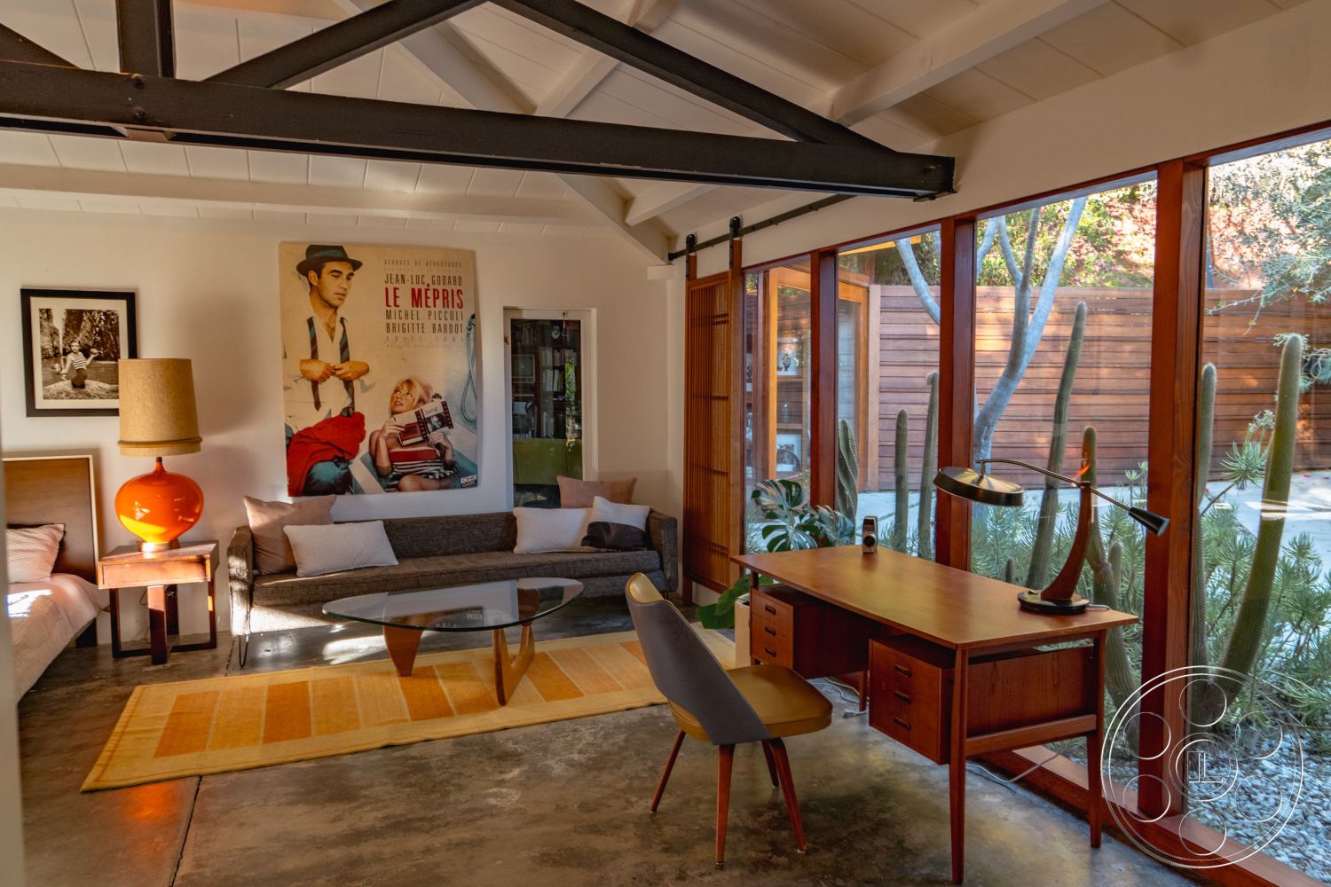 Pad 90210 - natural light interior, indoor plant decor interior, mid-century modern style interior, minimalist design interior, concrete floor interior, modern aesthetic interior, glass wall interior, round coffee table interior, textured wall art interior, exposed beam ceiling interior, wood frame windows interior, wooden desk interior, orange table lamp interior, soft seating area interior, cactus landscaping exterior, wooden fencing exterior, area rug accent interior