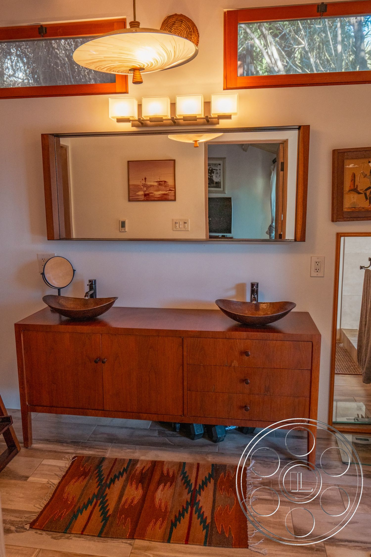 Pad 90210 - textured area rug, modern art wall decor, natural light from windows, overhead pendant lighting, neutral wall paint, minimalist bathroom design, contemporary bathroom decor, dual vessel sinks, wooden bathroom vanity cabinet, modern wall-mounted faucets, large framed mirror, wood trim around windows, planked wood flooring, open layout bathroom space, earth-toned color scheme, natural surroundings view