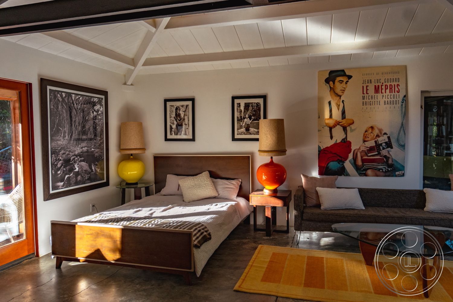 Pad 90210 - open floor plan, concrete flooring, framed black and white photographs, exposed wood ceiling beams, interior living space, mid-century modern design elements, natural light ambiance, wood nightstand with lamp, light-filled room, queen-sized bed frame with mattress, colorful ceramic table lamp, neutral upholstered throw pillows, vintage movie poster wall art, large glass windows with outdoor view, modern sectional sofa with cushions, abstract area rug with warm tones