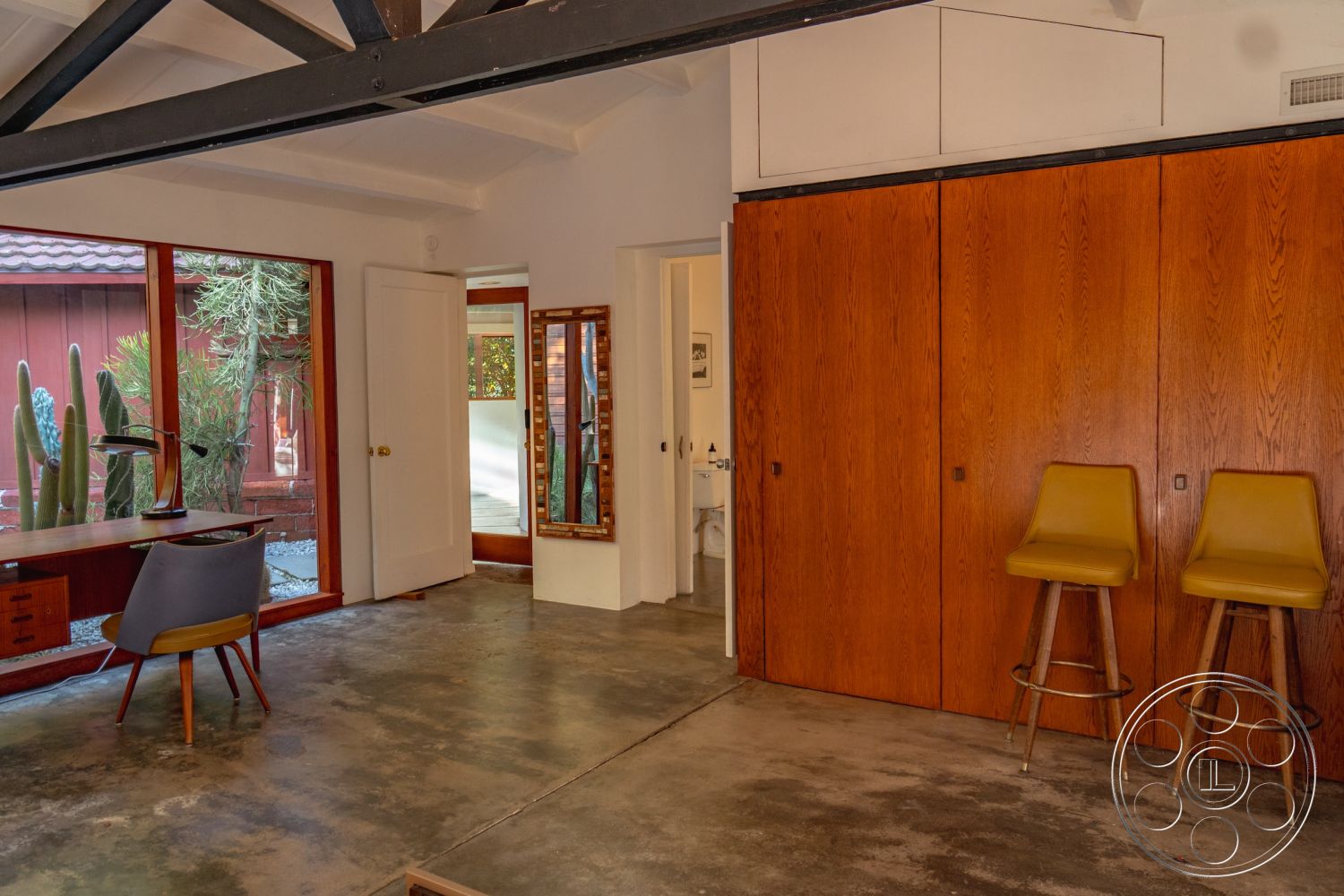 Pad 90210 - minimalist decor, open floor plan, sliding glass doors, greenery visible through windows, natural light from large windows, textured wall elements, mid-century modern design, exposed wood ceiling beams, suburban backyard view, contemporary art display, interior concrete floor, built-in wooden cabinets, wooden desk with ergonomic chair, yellow bar stools with wooden legs