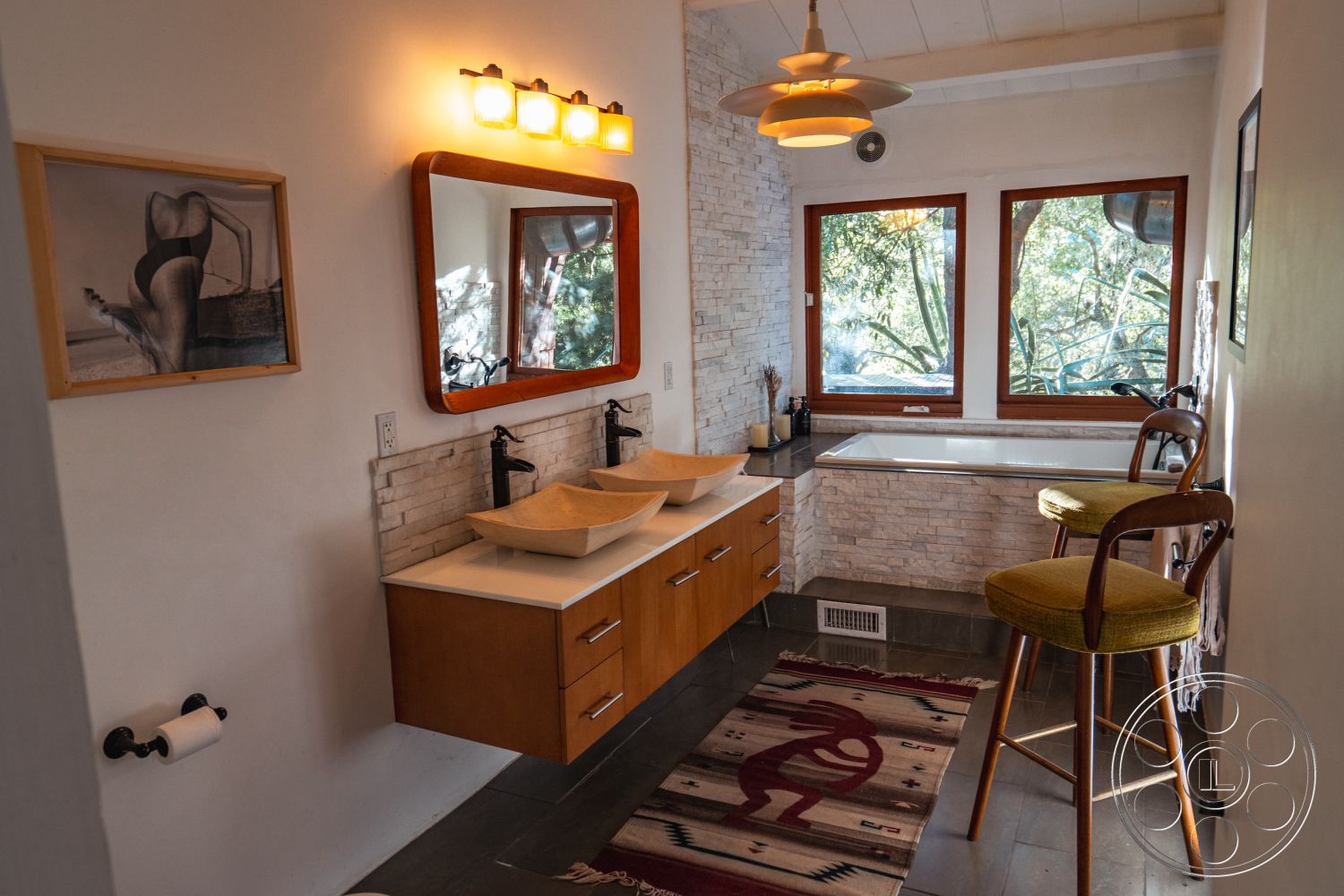 Pad 90210 - neutral color palette, soft ambient lighting, modern pendant lighting fixture, interior bathroom, textured stone accent wall, framed artwork on wall, natural stone wall treatment, wooden vanity cabinet with sink basins, contemporary round mirrors, gray slate flooring, large window views of greenery, built-in bathtub with stone surround, wooden bar stools with green upholstery, abstract patterned rug