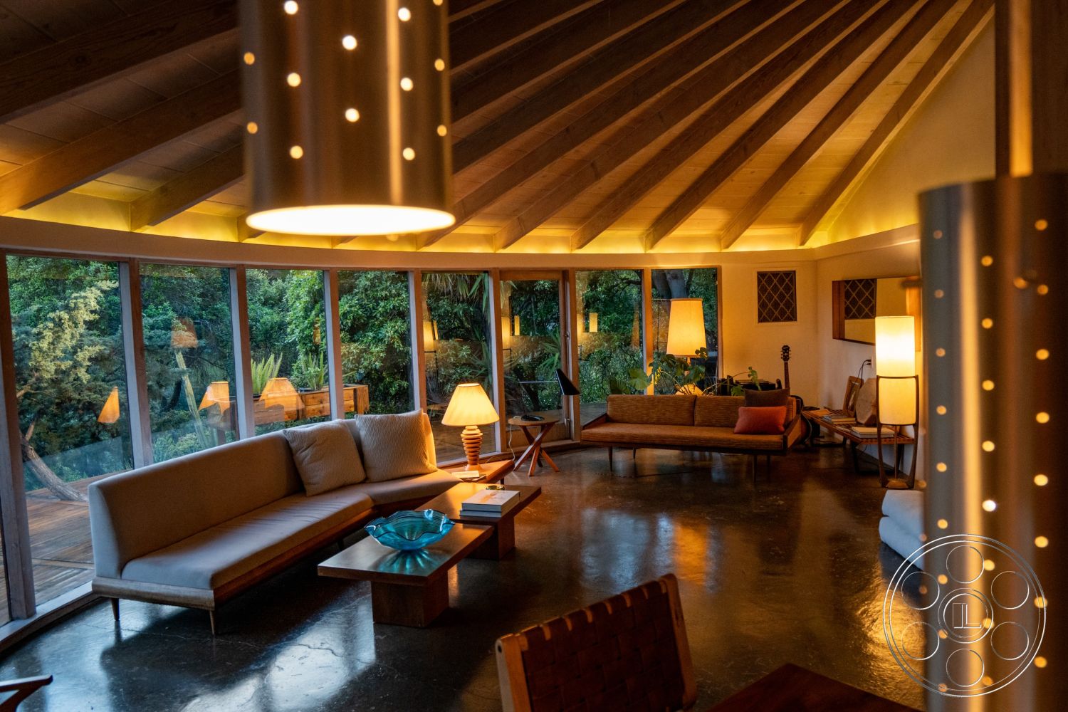 Pad 90210 - indoor plants, light wood furniture, round coffee table, cozy seating arrangement, open concept layout, textured wall panels, ambient lighting features, natural greenery view, large floor-to-ceiling windows, exposed wood ceiling beams, soft fabric sofa, minimalist decor style, vaulted living room ceiling, urban forest setting, mid-century modern interior, curved room layout, concrete floor finish in living room, glass decorative bowl, modern floor lamps, wooden deck view