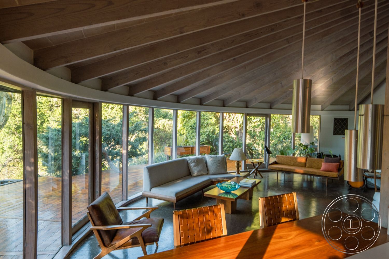 Pad 90210 - minimalist interior design, exposed wood ceiling beams interior, natural light-filled space interior, integrated indoor-outdoor living, open concept living area interior, polished concrete floor interior, curved glass walls interior, Mid-Century Modern architectural style, mid-century furniture design interior, wooden deck exterior, surrounding greenery view exterior, light-colored wood paneling ceiling interior, sitting area with geometric furniture interior, sculptural floor lamp interior, smooth, rounded furniture forms interior