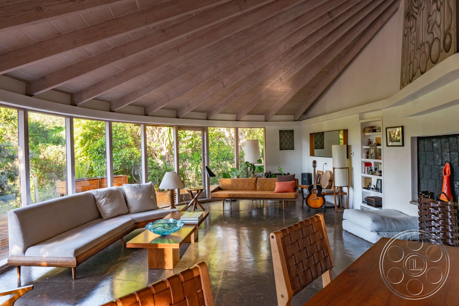 Pad 90210 - polished concrete flooring, glass coffee table, minimalist design elements, wooden dining table, artistic wall decorations, modern artistic style, earth-toned color palette, built-in bookshelves, comfortable and inviting space, exposed wood ceiling beams, integrated seating area, natural light flooding in, surrounding greenery view, interior open-concept space, curved glass wall windows, modern wooden sofa, wooden accents and decor, casual living room atmosphere, L-shaped layout, multi-functional living area, relaxed interior vibe