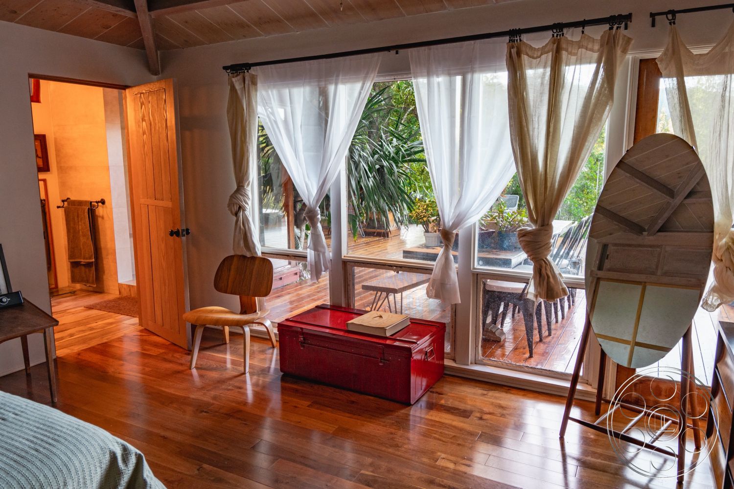 Pad 90210 - textured wall finish, natural light interior, contemporary interior style, open floor plan layout, hardwood bedroom floors, exposed wood beam ceiling, suburban backyard view, wooden deck patio, sheer curtain window treatments, glass panel windows, wooden interior doors, plastic garden furniture, rectangular red storage chest, modern chair design, indoor plant decorations, modern decorative mirror, bathroom with tile walls