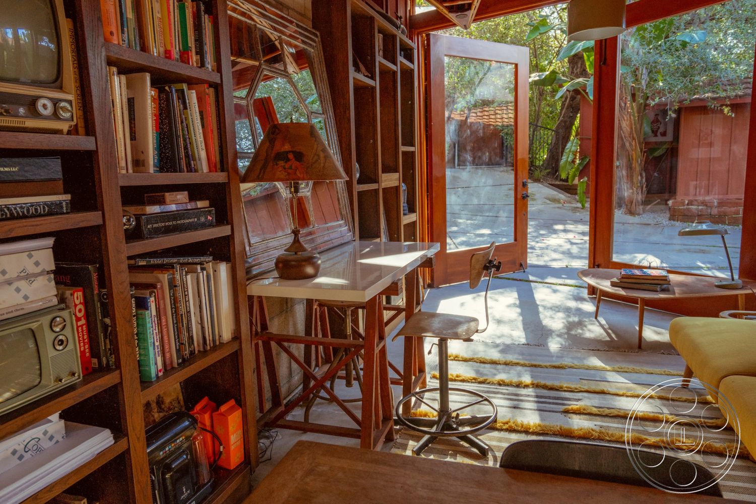 Pad 90210 - exposed wood ceiling beams interior, polished concrete floor interior, indoor plants for decor interior, natural light through large windows interior, wooden bookshelves with integrated lighting interior, glass sliding doors leading to outdoor area interior, mid-century modern style furniture interior, minimalist décor with artistic elements interior, striped area rug on floor interior, cozy reading nook with table lamp interior, simple desk workspace interior, open layout connecting indoor and outdoor spaces interior, harsh sunlight filtering through trees outside, wooden door frames accentuating warmth interior