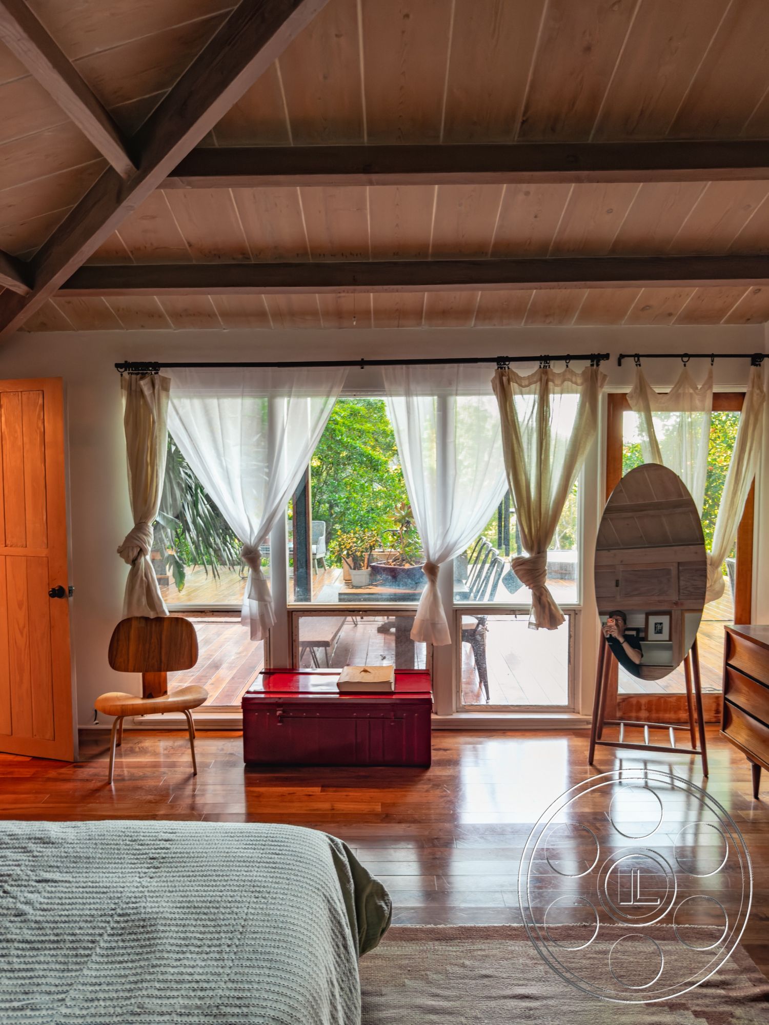 Pad 90210 - textured area rug, natural surroundings, floor-to-ceiling windows, modern minimalist decor, white sheer curtains, bright and airy space, contemporary armchair, exposed wood ceiling beams, hardwood bedroom floors, natural light-filled interior, red accent storage chest, wooden wardrobe door, outdoor deck view, relaxed bedroom atmosphere