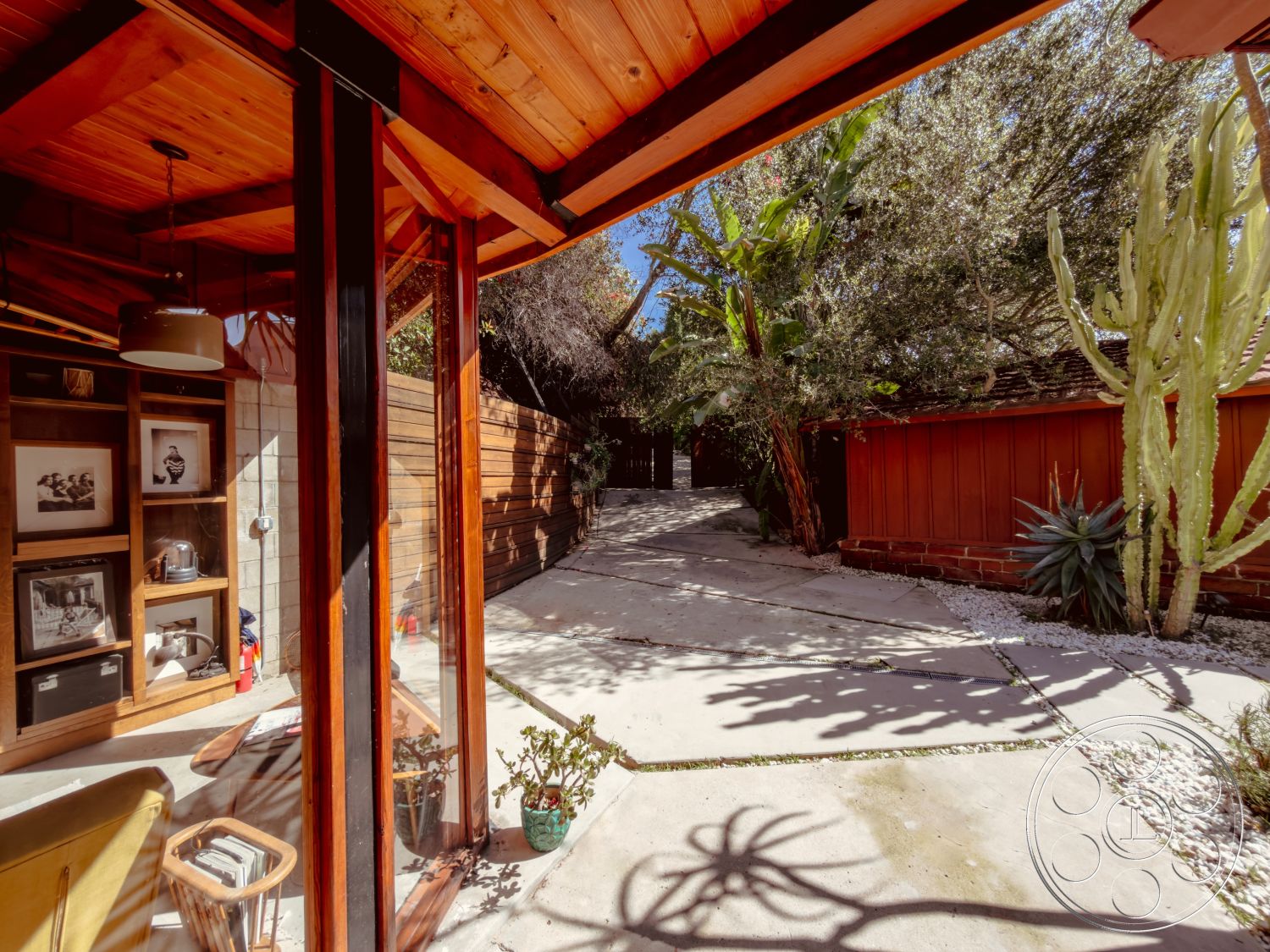 Pad 90210 - minimalist design elements, natural light from large windows, exposed wood ceiling beams, exterior concrete driveway, rectangular patio area, exterior concrete walkway, interior wood shelving units, textured stone wall treatment, glass wall panels, open layout space, wood beam architectural details, suburban backyard environment, landscape with desert plants, modern outdoor aesthetic, wooden fence structure, patterned tile accents, potted plants interior decor, architectural wooden accents