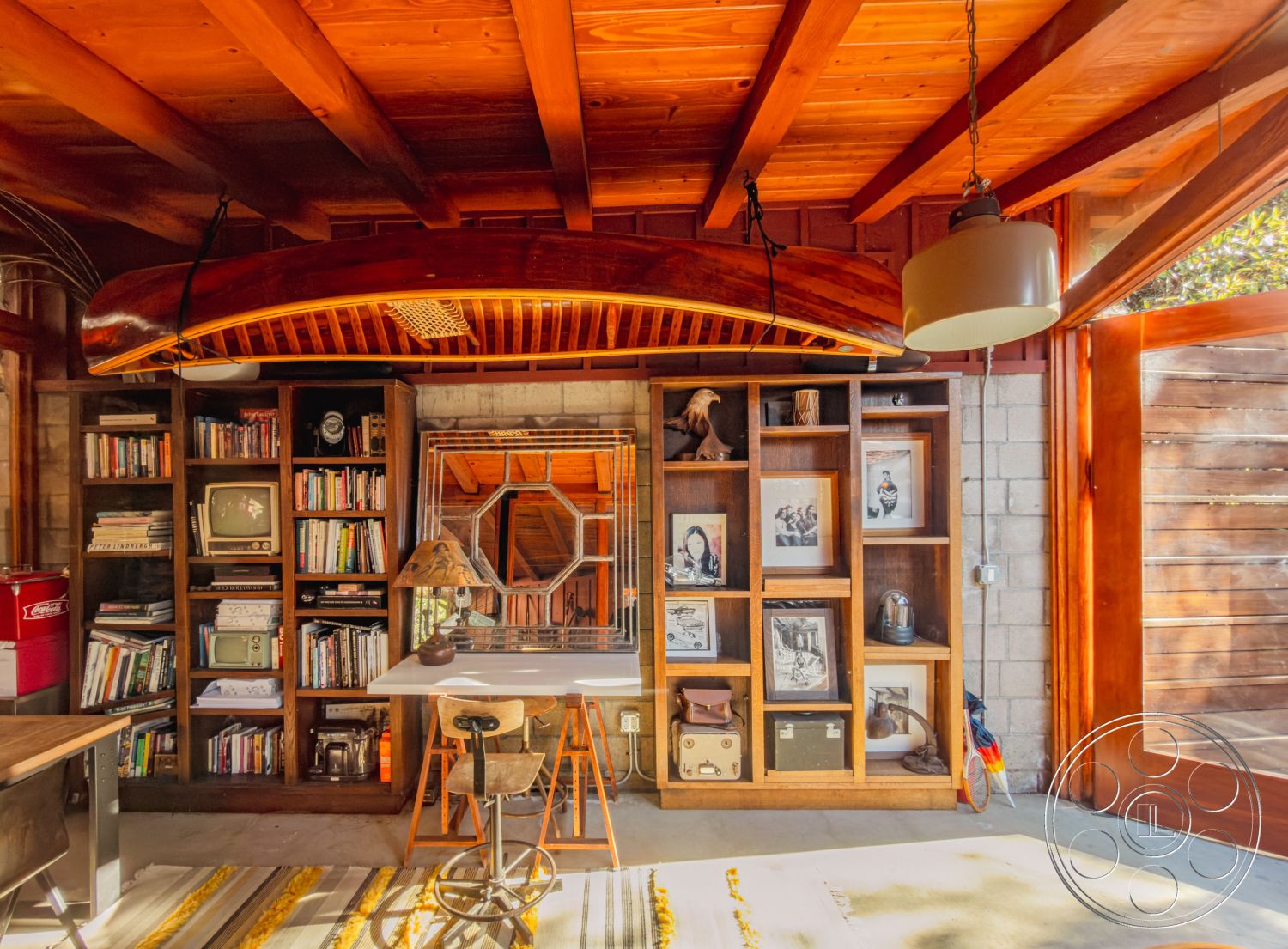 Pad 90210 - rug with geometric patterns, poured concrete flooring, modern rustic aesthetic, exposed wooden ceiling beams, glass paneled door, natural wood accent details, floating wood desk, custom built-in bookshelves, decorative plywood wall accents, minimalist industrial lighting, metal frame chair, hexagonal mirror decoration, console table with vintage decor, study area with coastal decor, artistic display of vintage items, interior workspace with natural light