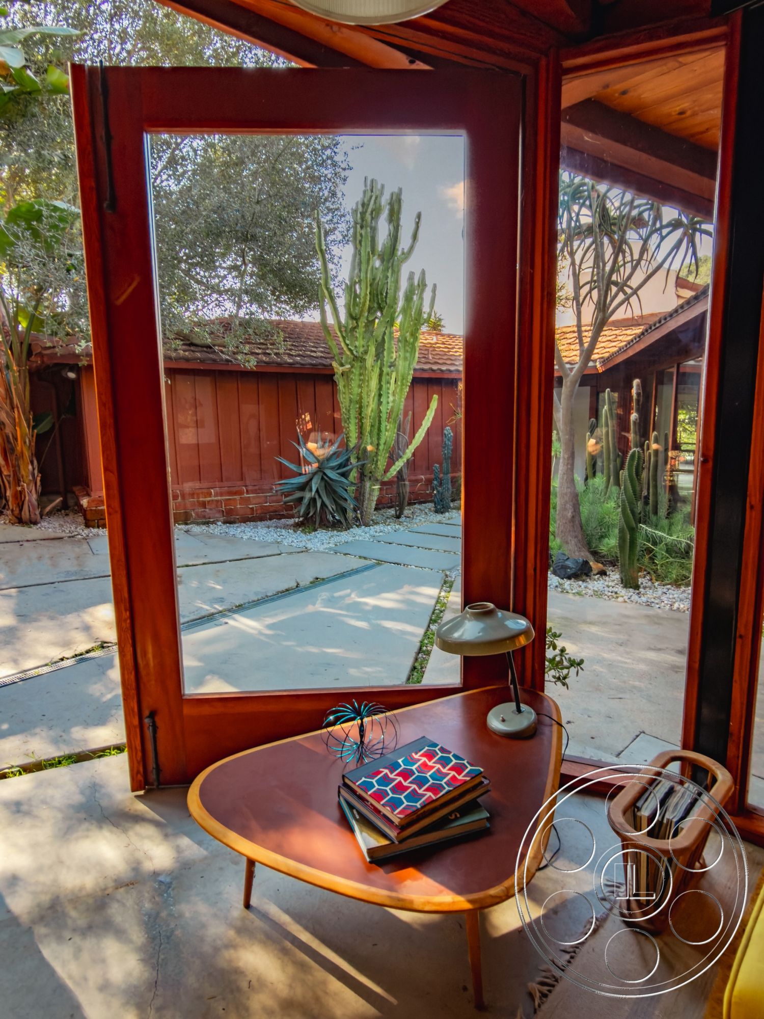 Pad 90210 - interior vaulted ceiling, interior natural light, interior cozy reading nook, interior minimalist design, interior concrete floor, interior wood framed windows, exterior desert landscaping, interior triangular coffee table, interior modern lamp, interior stacked books, exterior cacti garden, exterior gravel pathways, exterior red wooden siding, interior organic decor, exterior clear sky view, exterior Mediterranean style