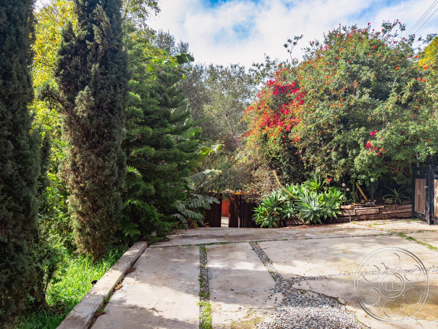 Pad 90210 - lush tropical landscaping, natural stone planters, exterior concrete driveway, exterior lush greenery, sunlit outdoor setting, urban garden environment, exterior gravel pathway, exterior flowering shrubs, exterior tall conifer trees, exterior wooden fencing, textured stone retaining wall, inviting outdoor entrance, curved driveway with grass borders