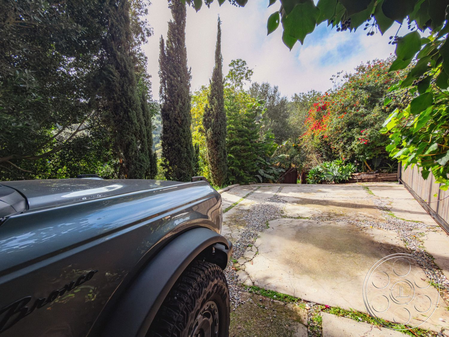 Pad 90210 - mountain view, surrounded by greenery, tall cypress trees, semi-rural location, natural stone retaining wall, rustic outdoor setting, lush garden landscaping, exposed aggregate concrete surface, seasonal flowering plants, exterior gravel driveway, round stone planters, patio area with natural vegetation