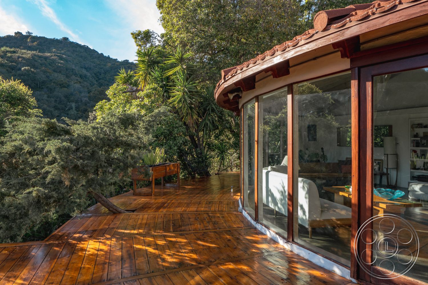 Pad 90210 - natural wood flooring, natural wood railing, contemporary architectural style, mountain view setting, exterior wooden deck, surrounding lush greenery, scenic forest backdrop, open floor plan living area, red clay roof tiles, sunlit outdoor area, transitional indoor-outdoor space, curved glass walls, wood beam design elements, integrated planter box, rustic outdoor entertaining space