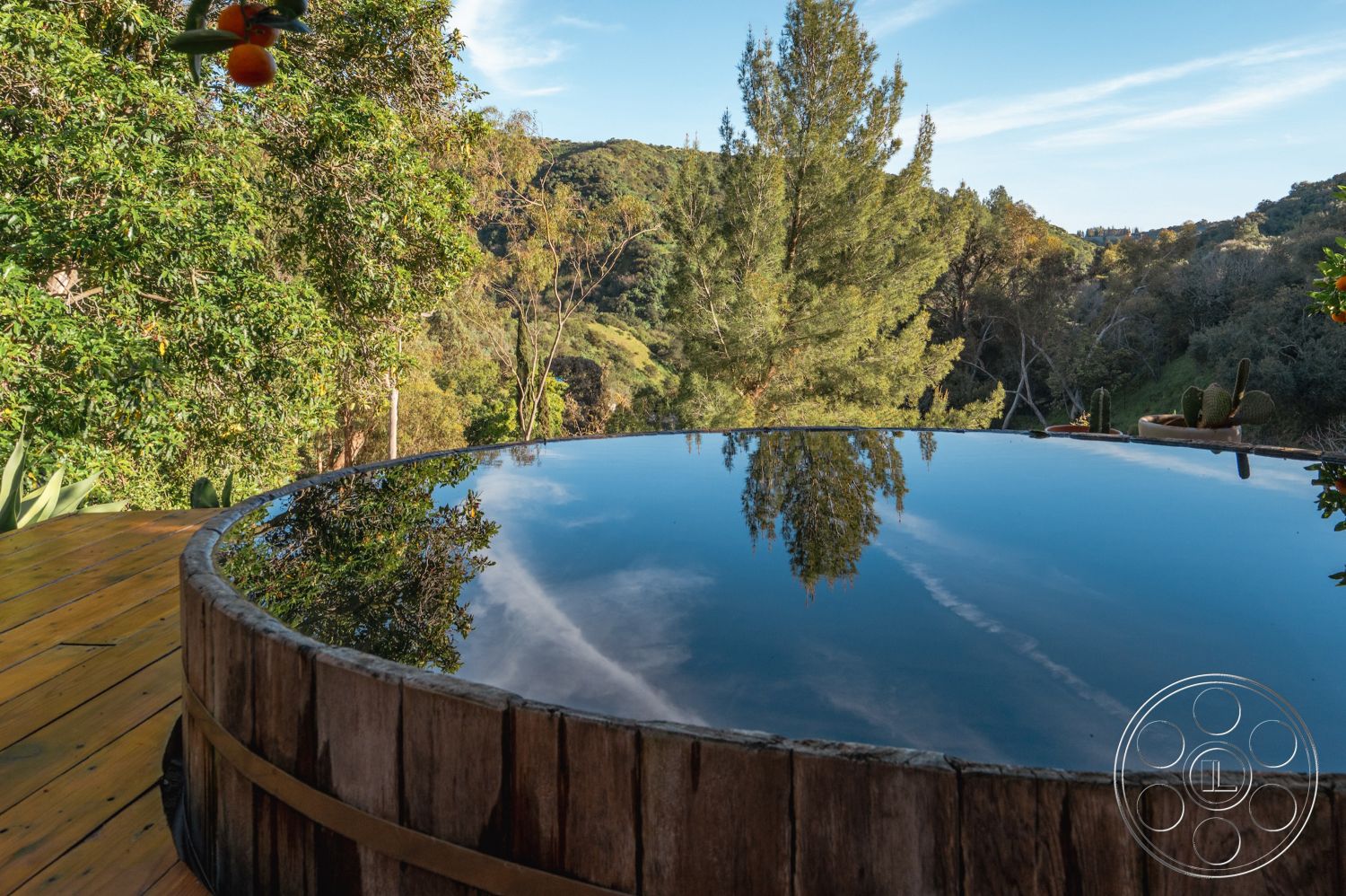 Pad 90210 - lush greenery, sloped terrain, circular hot tub, outdoor living space, mountain view, reflective water surface, wooden hot tub, surrounding forest setting, natural landscape backdrop, exterior relaxation area, natural wood deck flooring, wooden railing details