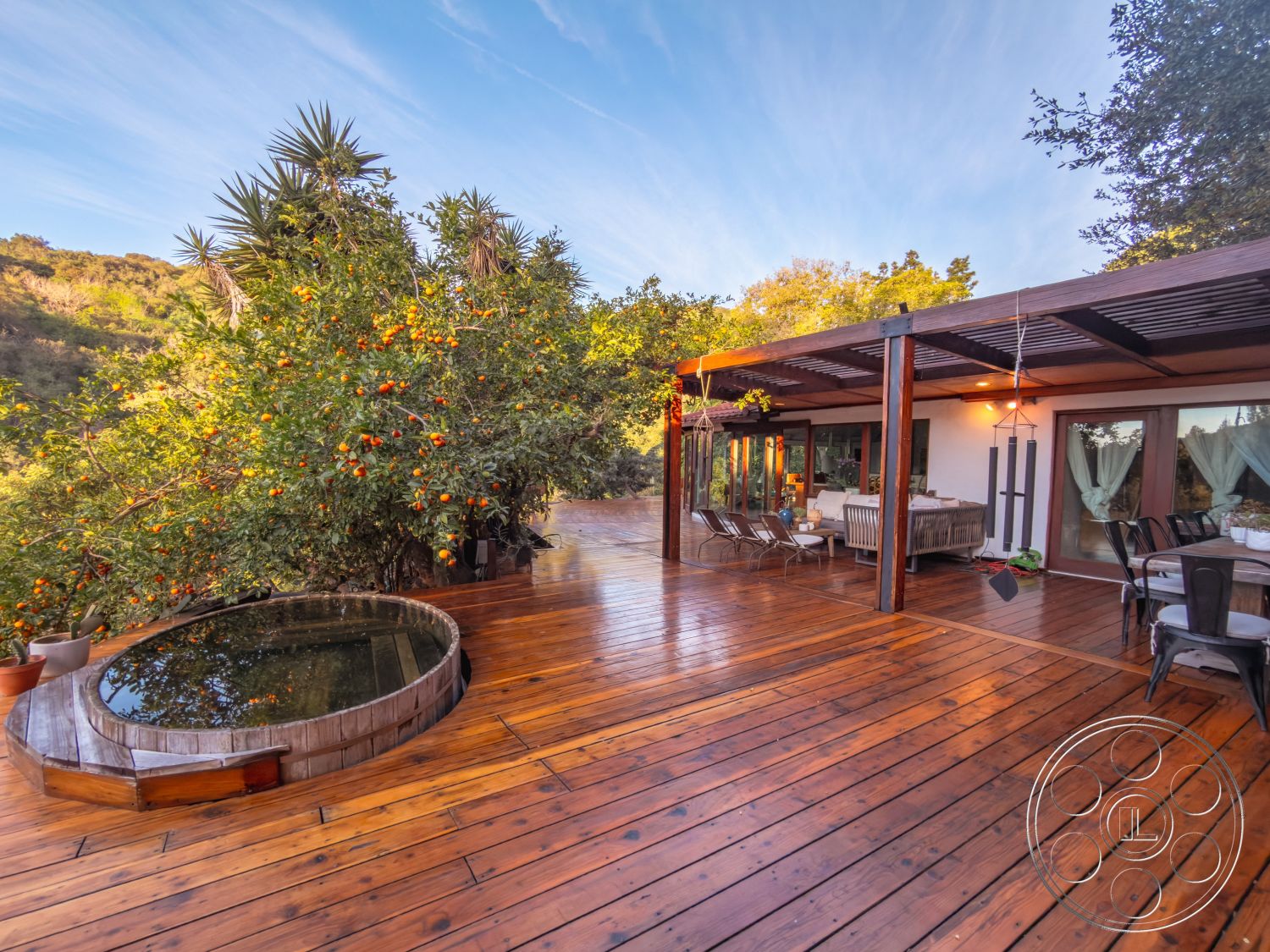 Pad 90210 - city skyline view, open concept living space, glass patio doors, lush garden setting, modern outdoor seating area, exterior wooden deck flooring, wooden pergola structure, string lights for ambiance, round wooden hot tub, curved deck design, potted citrus trees, natural landscape background, rustic outdoor dining table, slender wooden support beams