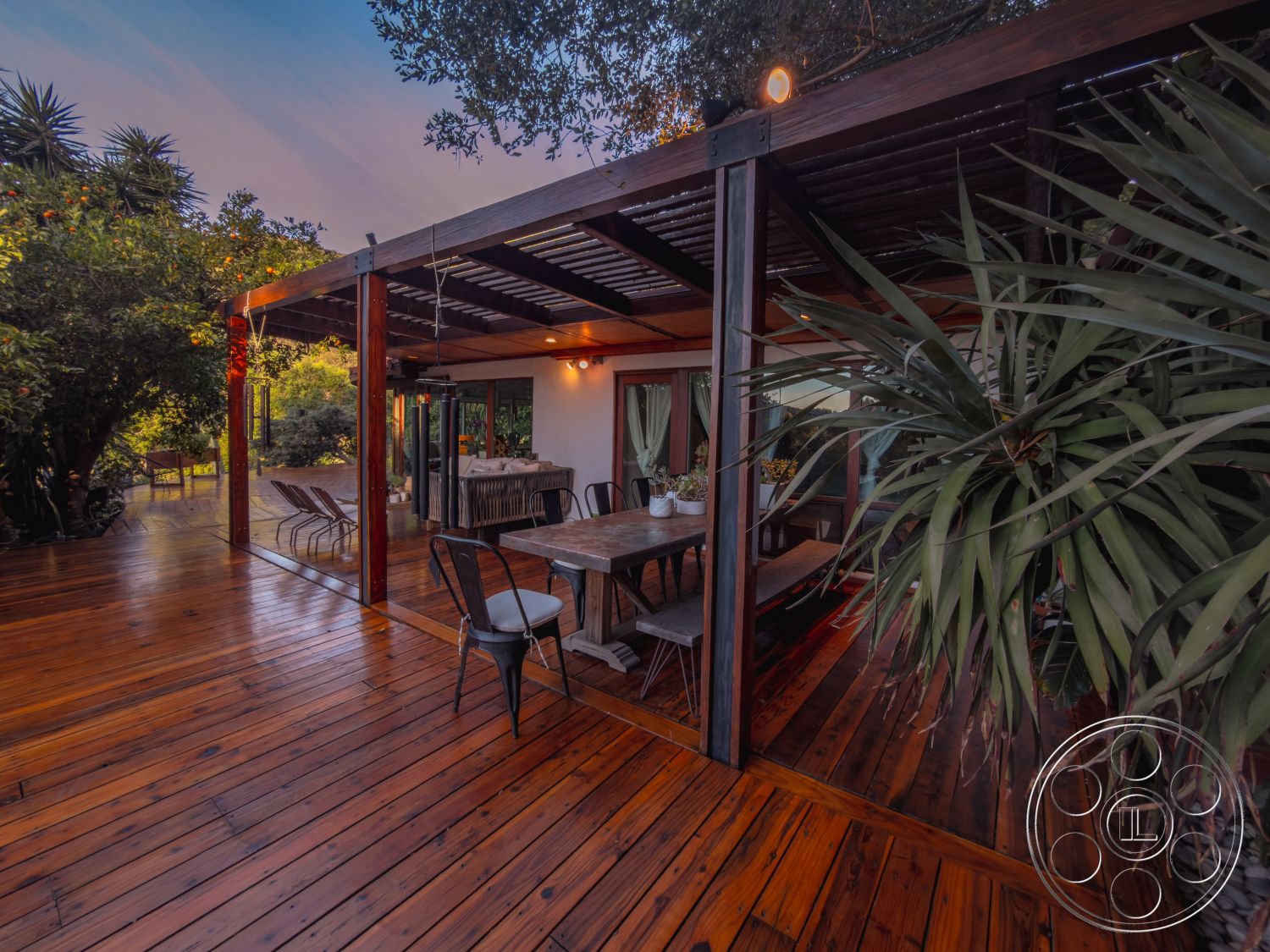 Pad 90210 - natural wood dining table, sunset view, outdoor lighting fixtures, modern outdoor furnishings, landscaped garden area, surrounding lush greenery, exposed wooden ceiling beams, exterior wooden deck flooring, open-air dining area, relaxed seating arrangement, private backyard setting, pergola with metal accents, potted plants on dining table, contemporary black metal chairs, freestanding structure with glass walls, rustic charm outdoor space, inviting outdoor atmosphere