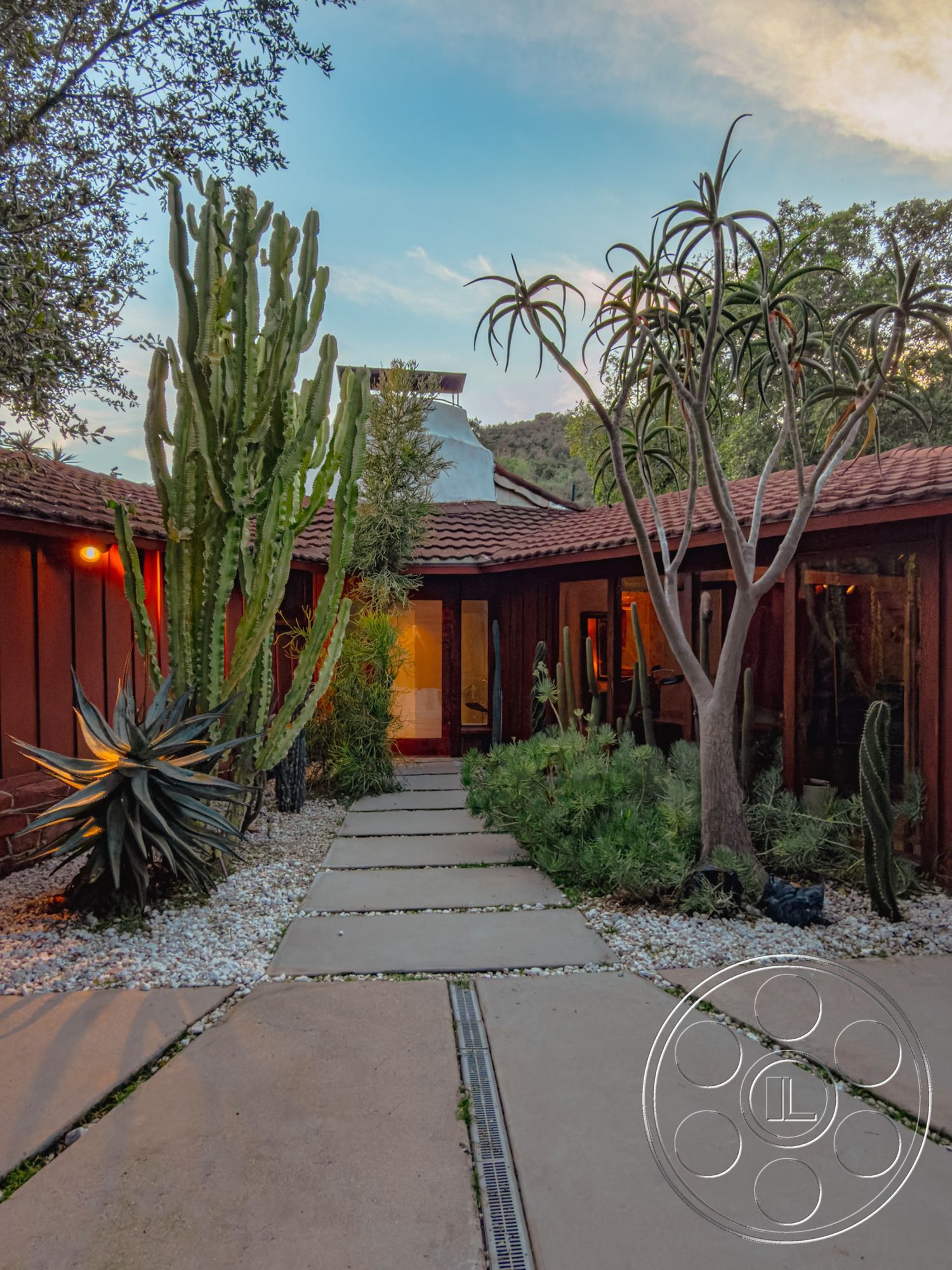 Pad 90210 - low-maintenance garden, desert landscaping, red tile roof, wooden wall panels, drought-resistant plants, cacti garden, native shrubs, glass door entrance, exterior concrete pathway, open-air patio area, flat concrete pavers, courtyard-style home, surrounding hill view, warm evening light