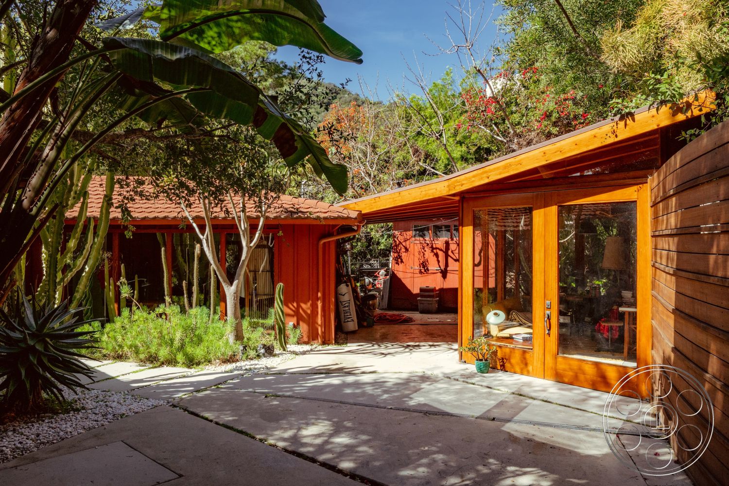 Pad 90210 - open floor plan, sliding glass door, natural wood finishes, modern outdoor space, angled roofline, desert-inspired landscaping, flat roof design, landscaped garden area, concrete patio flooring, urban garden setting, minimalist outdoor furniture, glass door entrance, curved stone pathway, exterior wooden siding, natural light-filled interiors, wooden deck entryway, red painted exterior walls, tropical plants surrounding property, brightly colored exterior accents